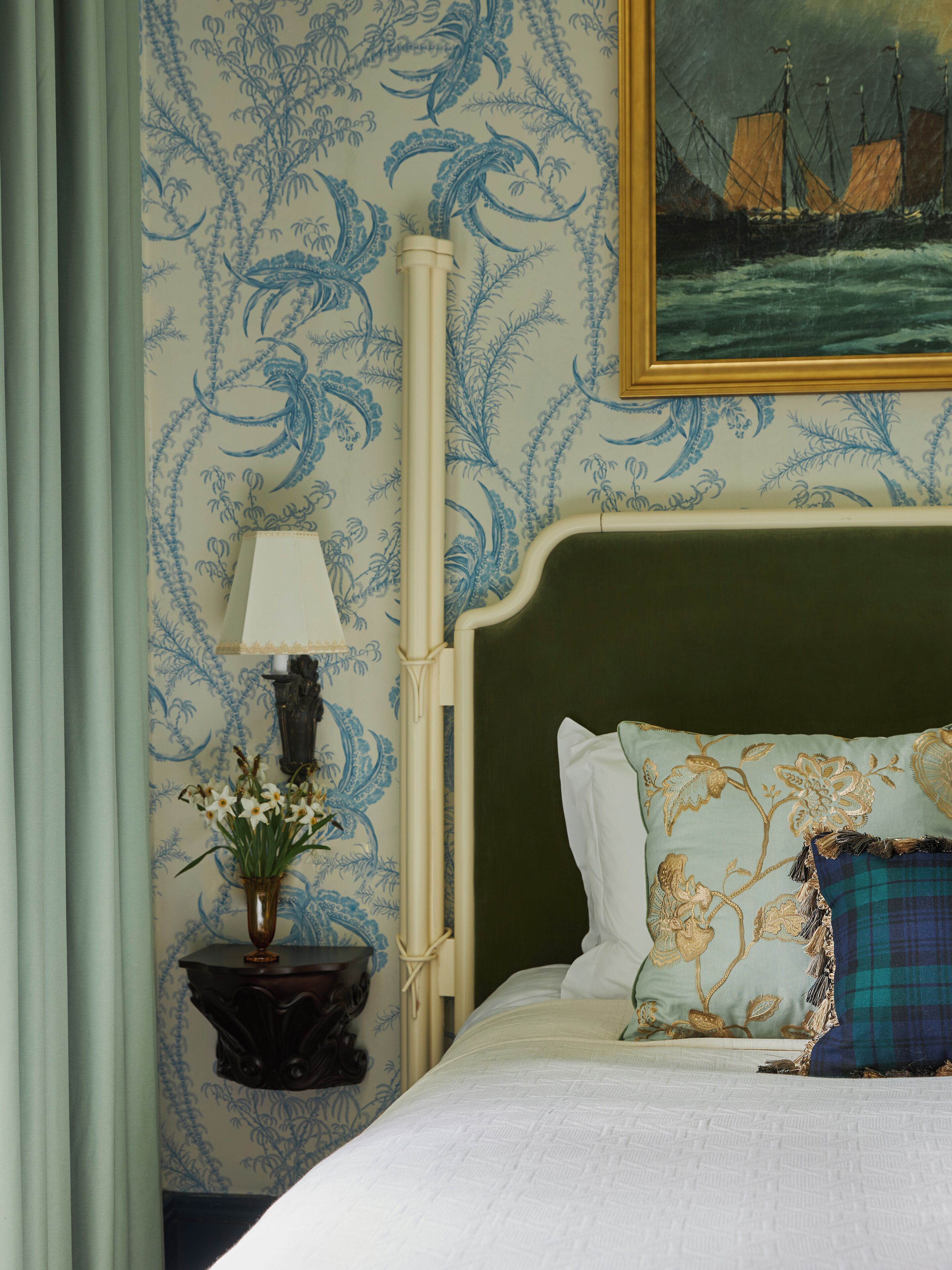 A neatly made bed with floral pillows, a bedside table with a lamp and flowers, and a wall with a maritime painting.
