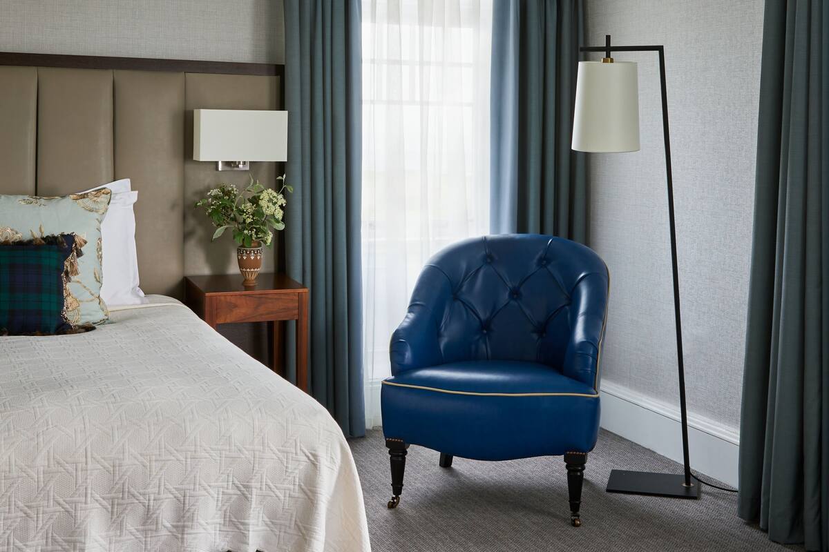 A hotel room with a bed, a blue armchair, a floor lamp, and a window with curtains.