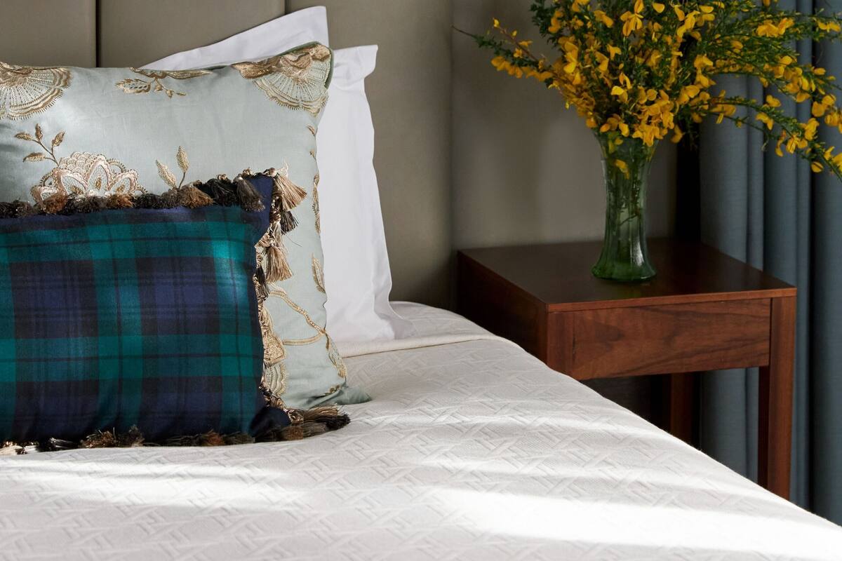 A neatly made bed with a white and teal plaid pillow, a white pillow, and a vase with yellow flowers on a bedside table.