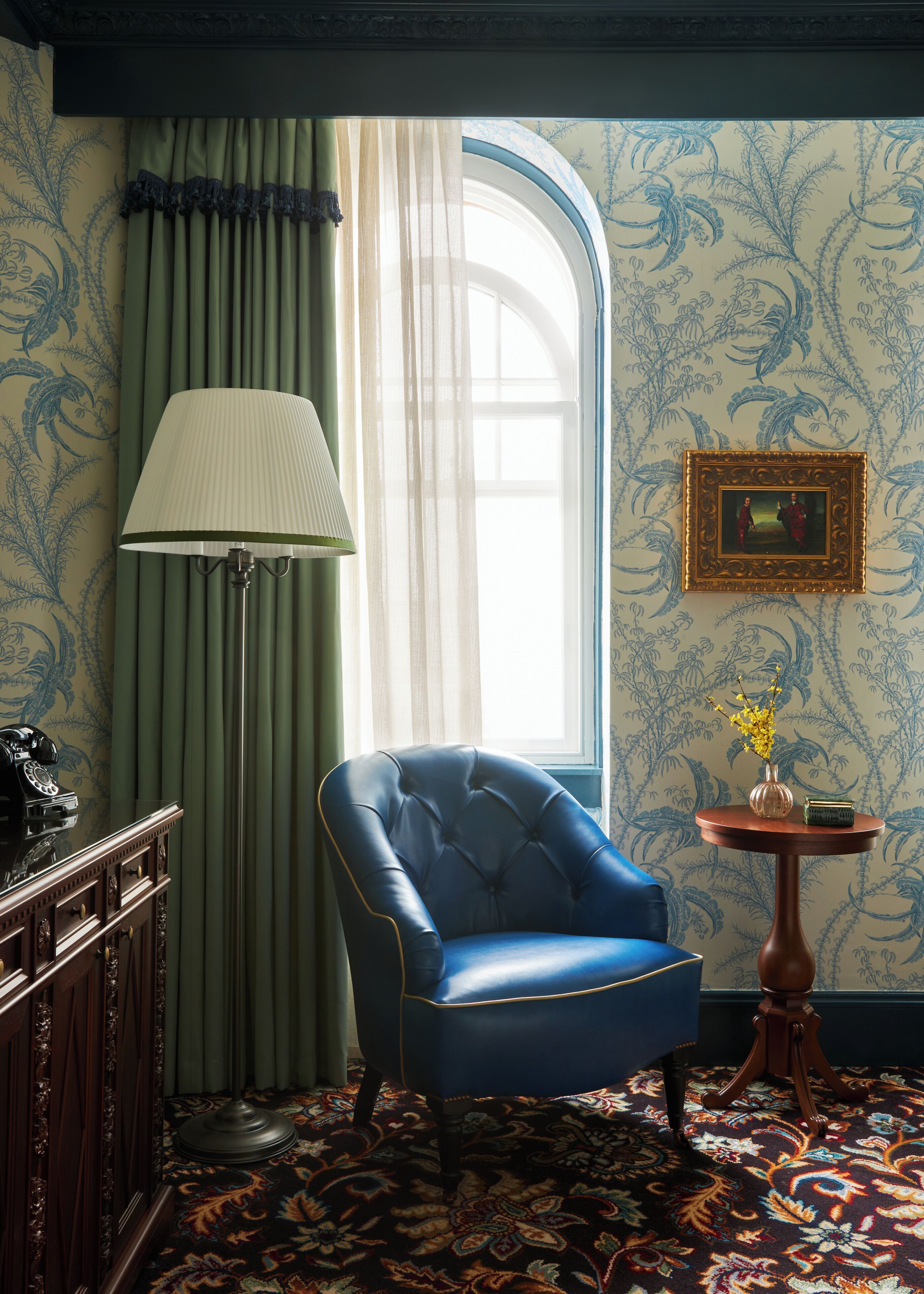 Um quarto com uma poltrona azul, uma luminária, um telefone, uma mesinha de centro com um vaso, e um quadro pendurado na parede.