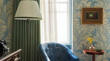 Um quarto com uma poltrona azul, uma luminária, um telefone, uma mesinha de centro com um vaso, e um quadro pendurado na parede.