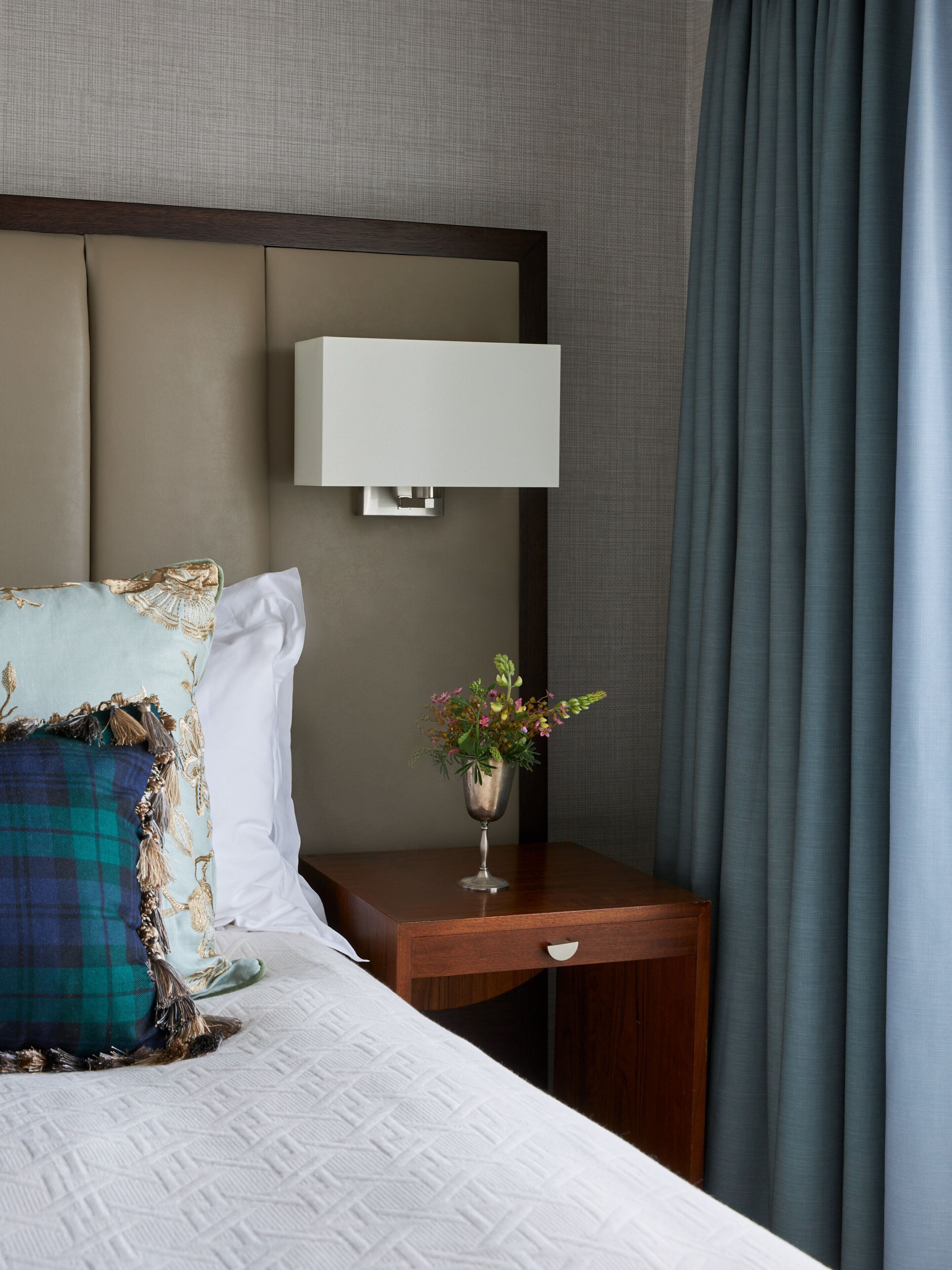 Un lit bien fait, avec une couette blanche et des coussins décoratifs, une table de chevet ornée d’un vase de fleurs et une lampe fixée au mur.