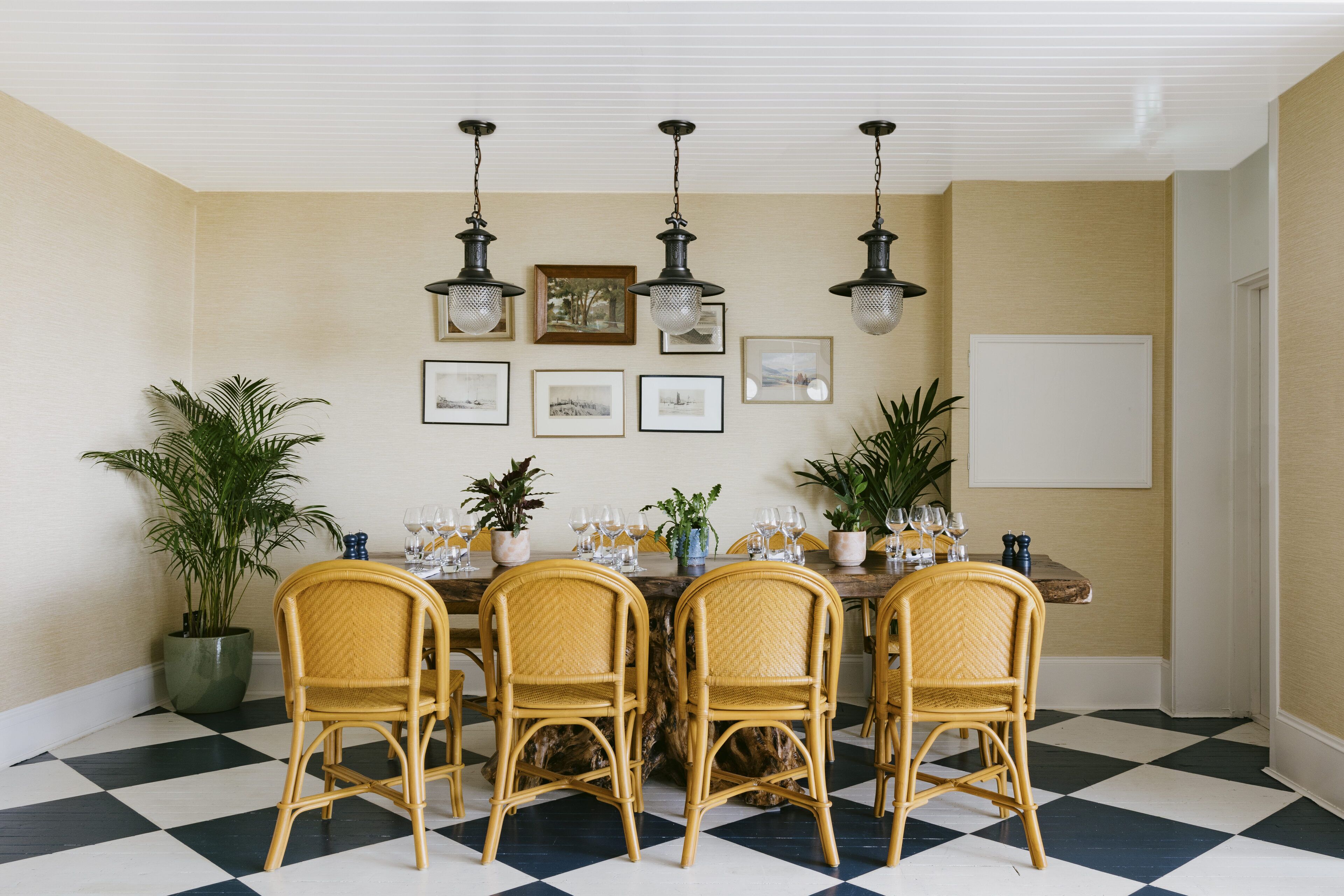 A dining area with a checkered floor, four wicker chairs, a wooden table set with glasses and a centerpiece, and wall-mounted pendant lights.