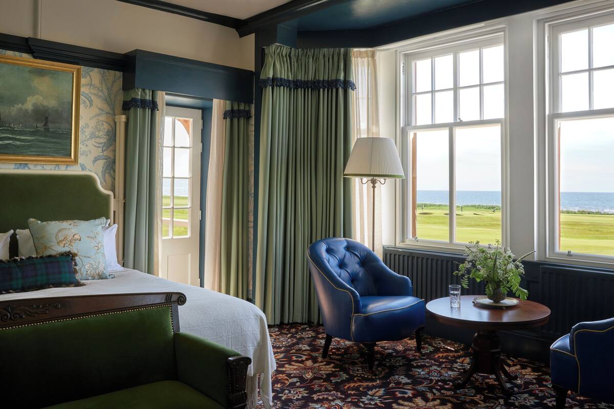 A hotel room with a bed, a chair, a table with a plant, and a view of the sea through large windows.