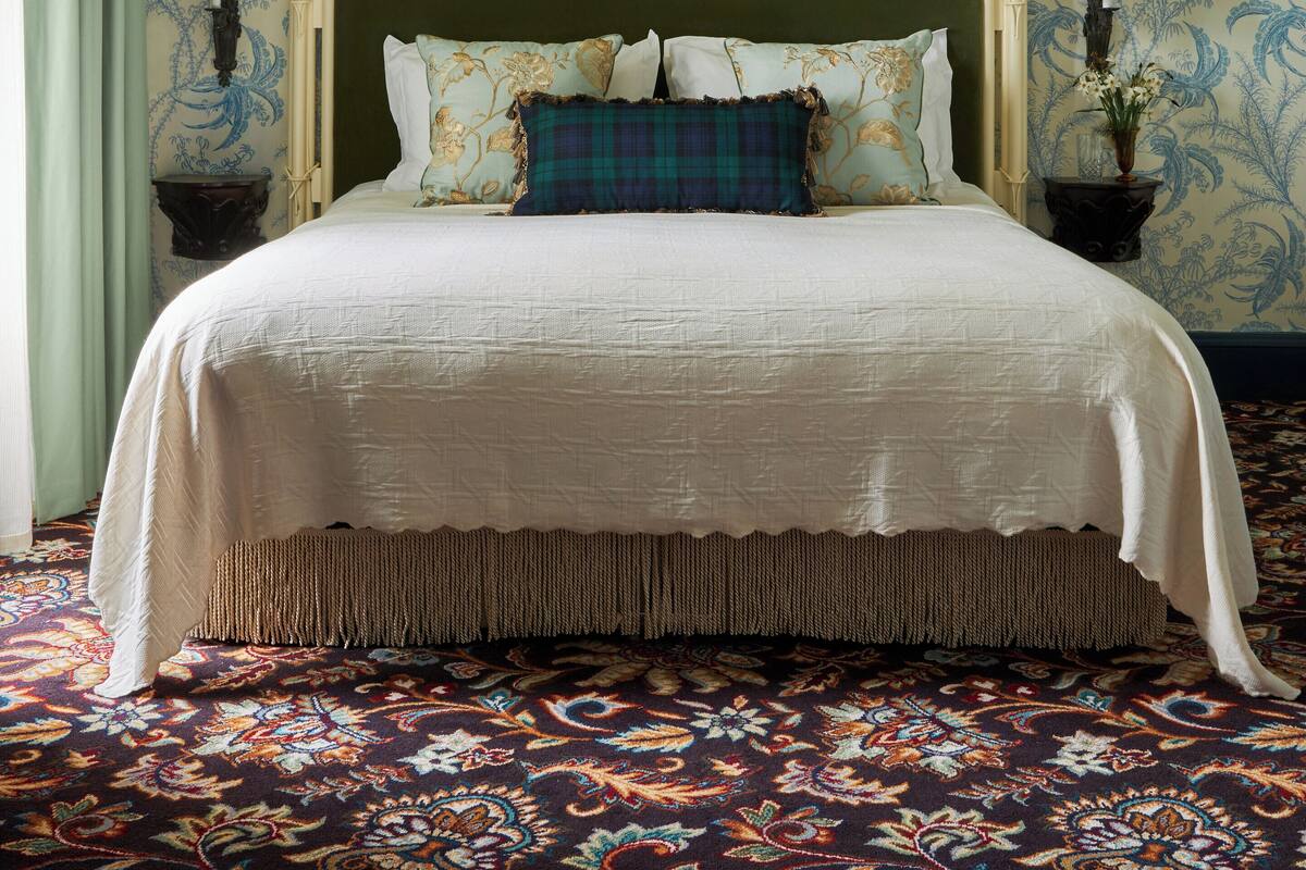 A bed with a green headboard, white bedding, and a plaid pillow. There is a painting above the bed and two bedside tables with lamps.