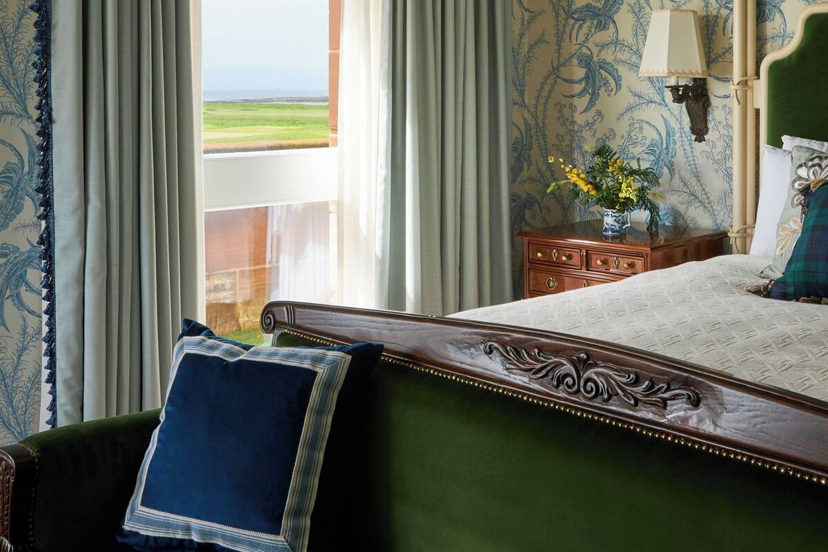 A room with a bed, a green upholstered chair, a wooden nightstand, and a view of the outdoors through a window with curtains.