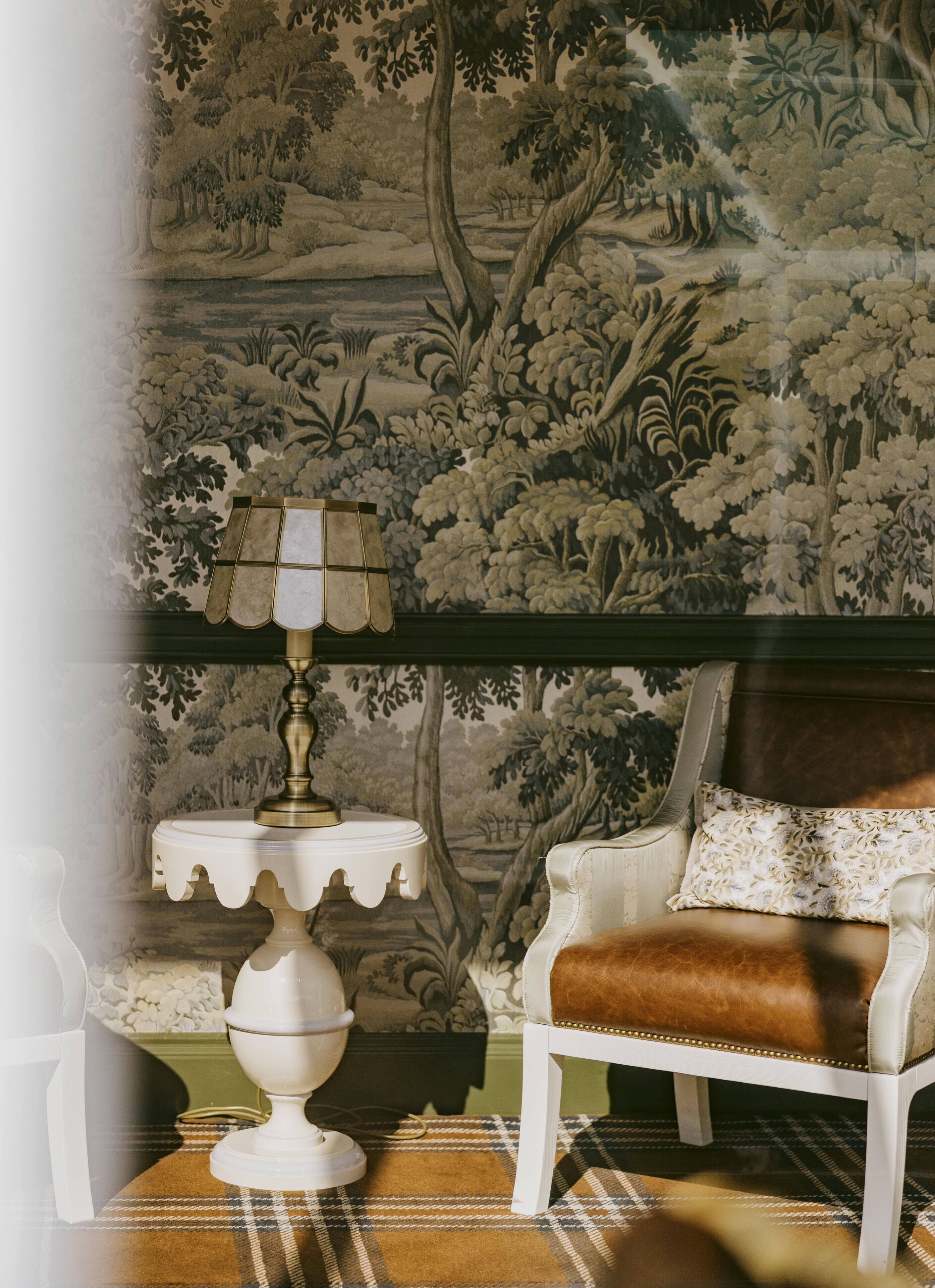 A room with a patterned wallpaper, a white chair with a brown leather seat, a white side table with a lamp, and a plaid rug.