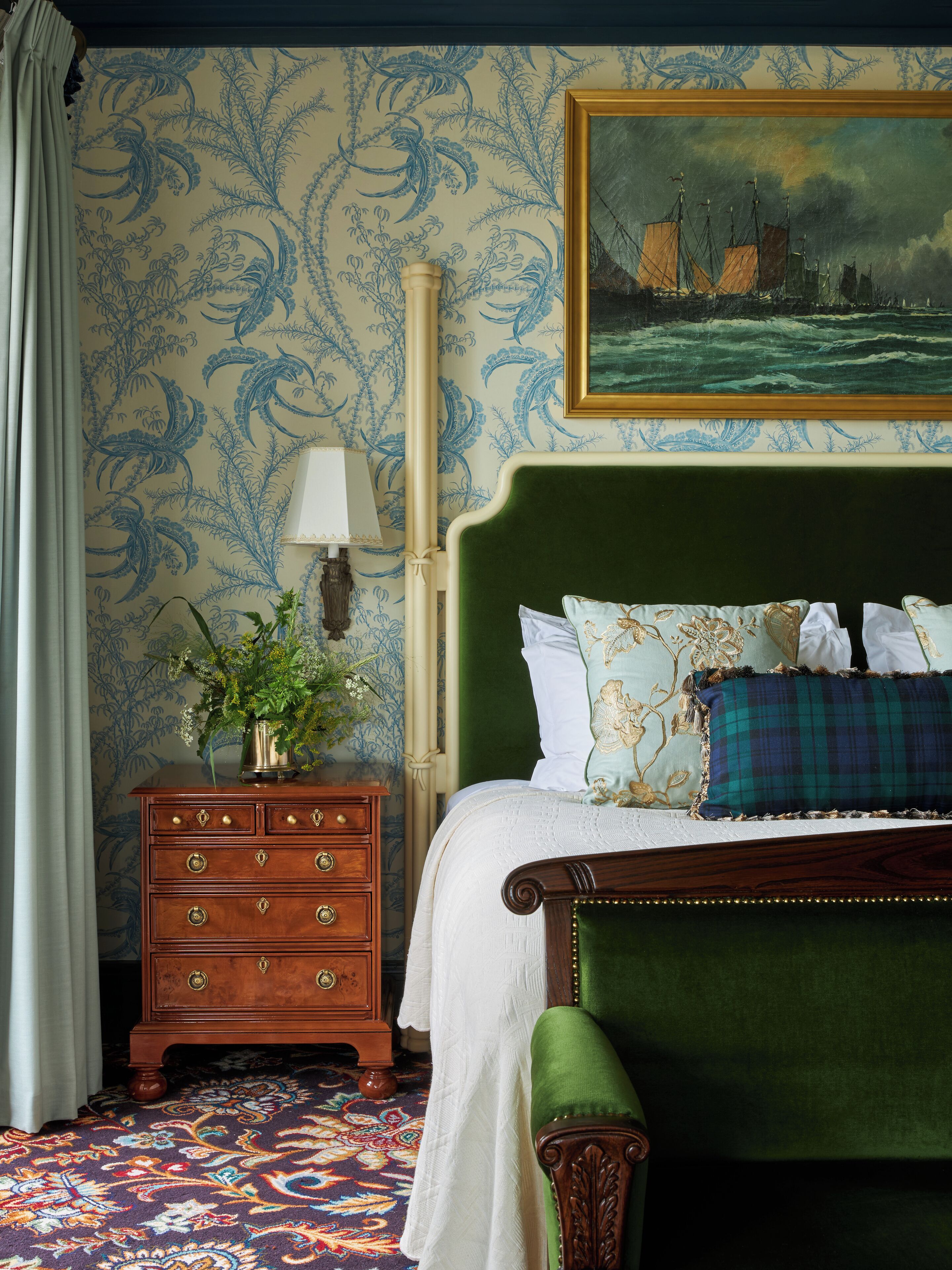 Un dormitorio con una cama tapizada de verde, una mesita de noche de madera con cajones, una lámpara y un papel tapiz estampado con motivos florales.