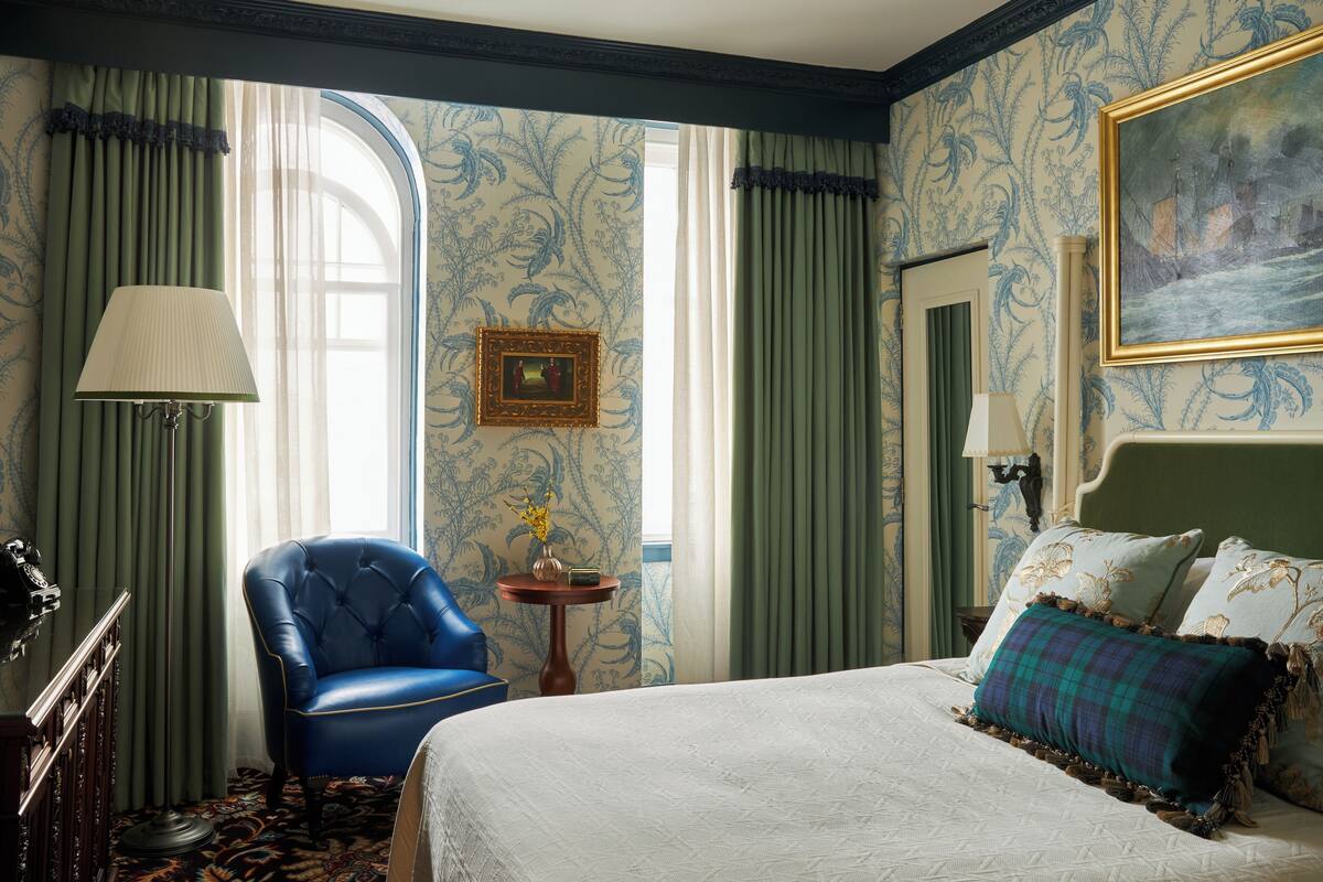 A hotel room with a large bed, a blue armchair, a small table with a vase, and a painting on the wall.