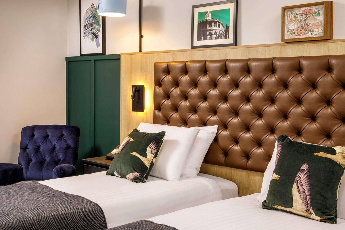 Habitación de hotel con un cabecero capitonado, dos camas con sábanas blancas, un sillón azul oscuro, un armario verde y obras de arte enmarcadas en la pared.