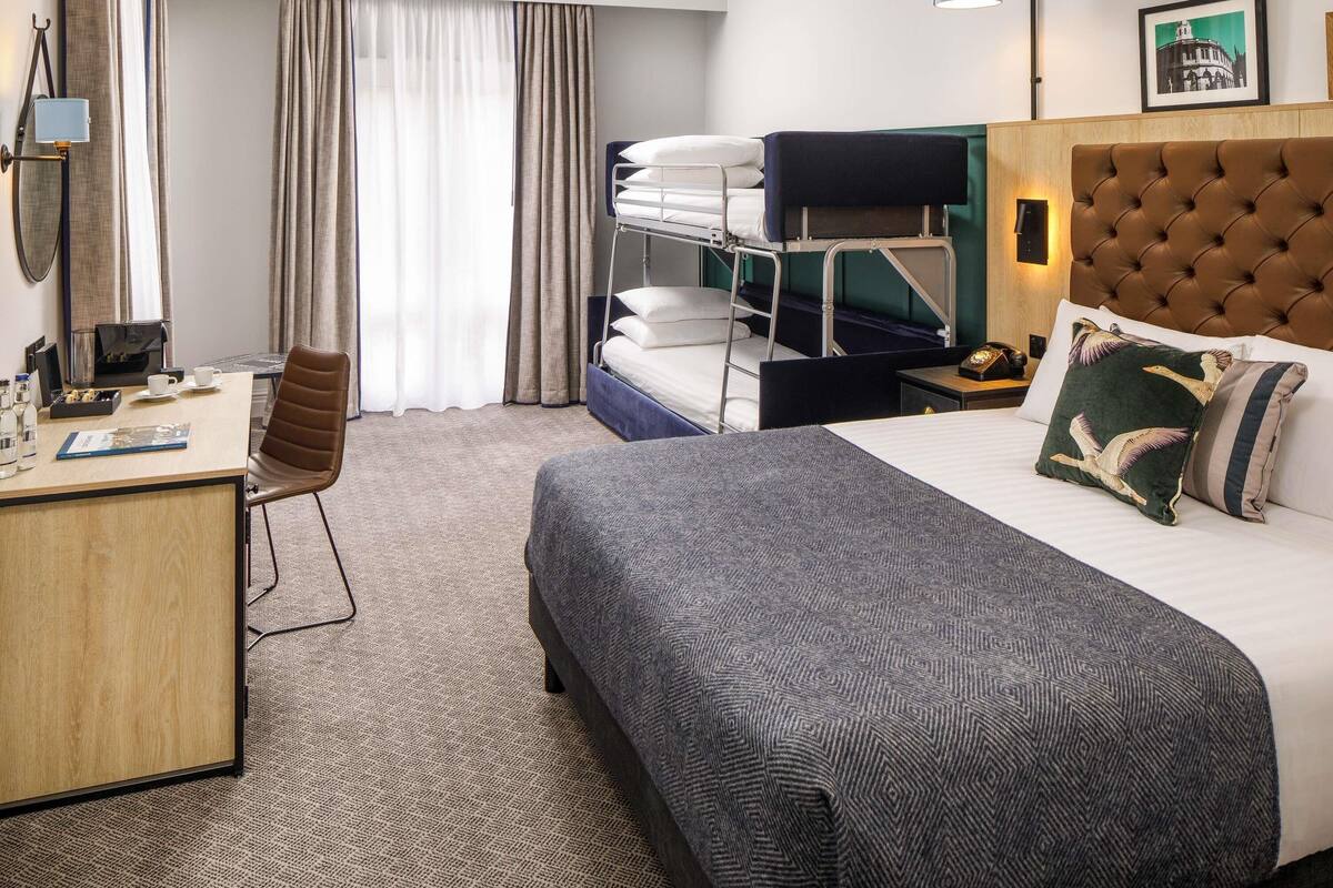 Una habitación de hotel con literas, un escritorio con una silla, una mesita de noche con lámpara y un cuadro en la pared.