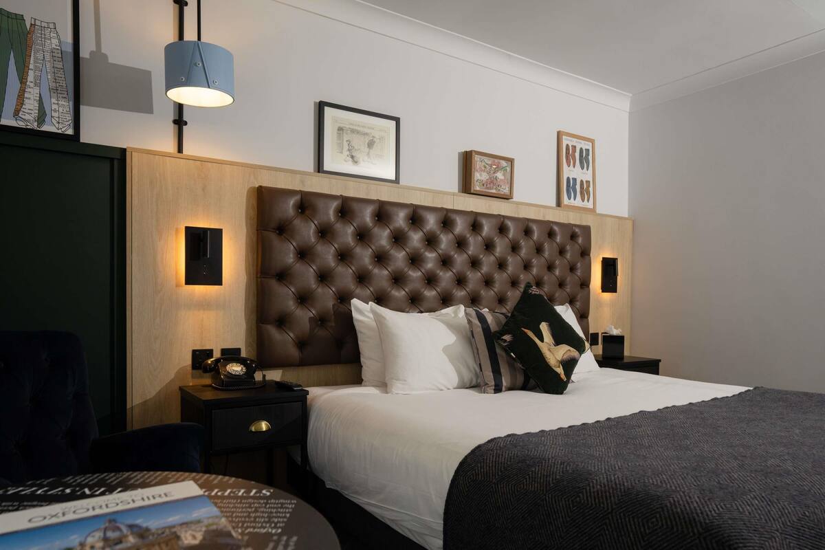 Habitación de hotel con un cabecero capitonado, una mesita de noche redonda con lámpara, cama con ropa de cama blanca y un cuadro enmarcado sobre el cabecero.