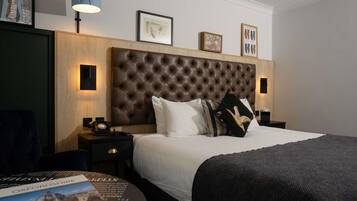A hotel room with a tufted headboard, a round bedside table with a lamp, a bed with white linens, and a framed picture above the headboard.