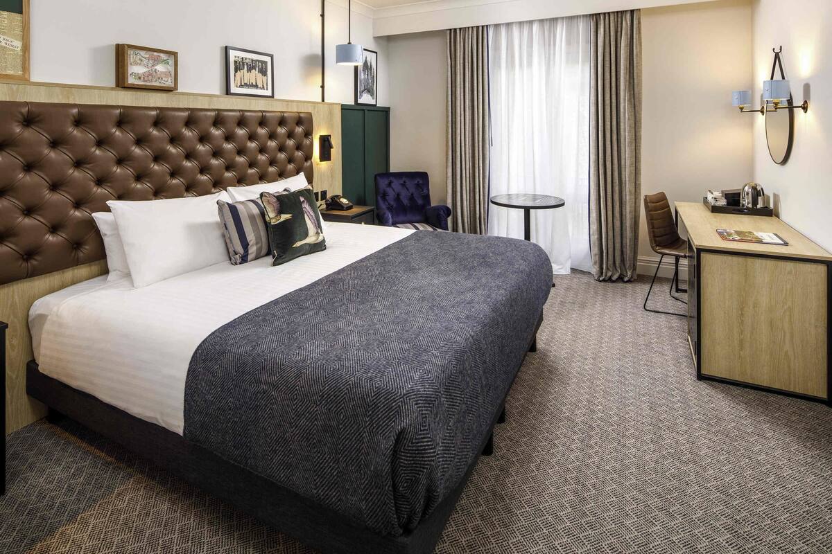 Habitación de hotel con una cama grande, un escritorio, una silla, una mesita y una lámpara de pared.