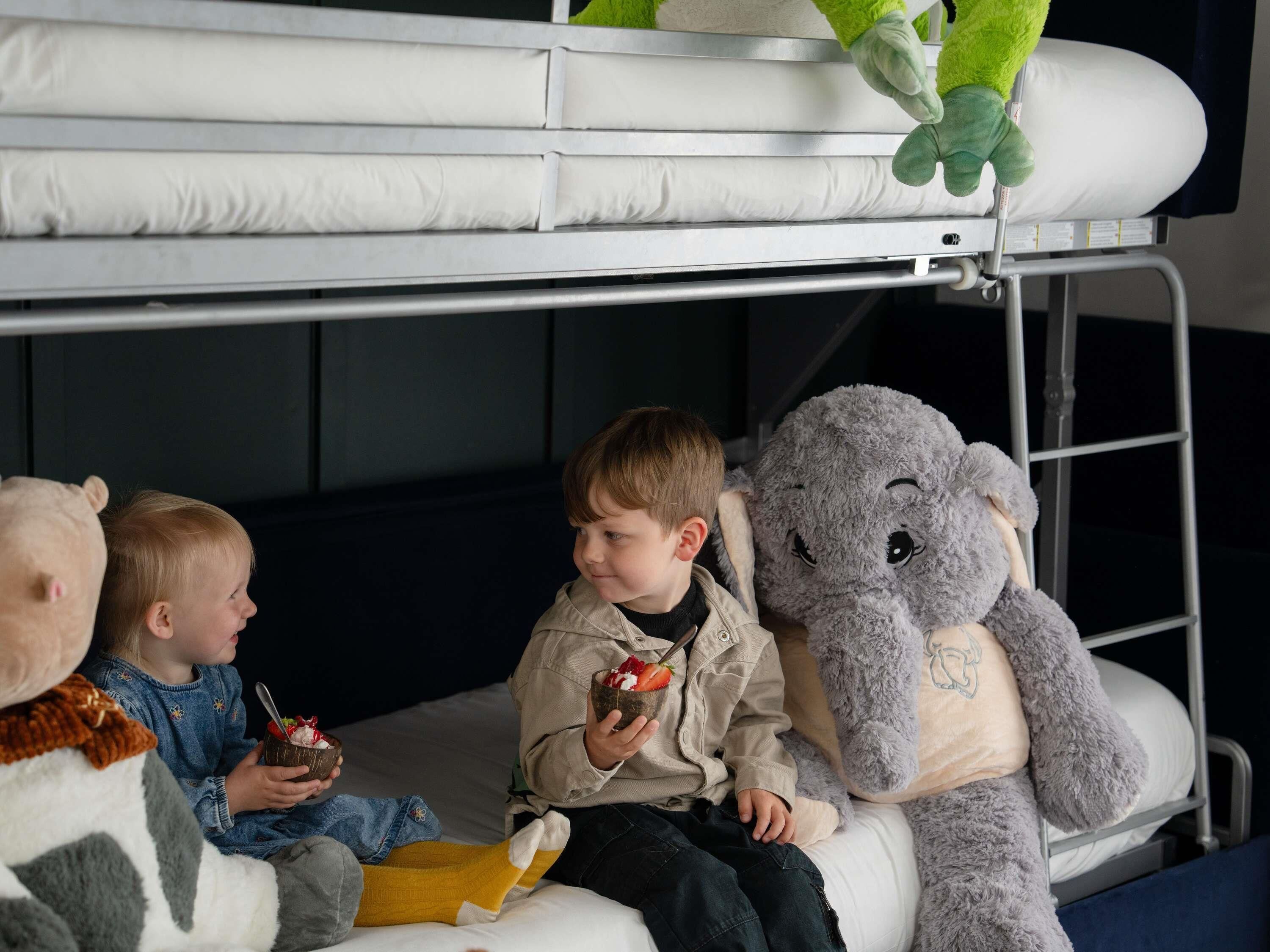 A bunk bed with two children sitting on it, one holding a strawberry, and a large stuffed elephant toy beside them.