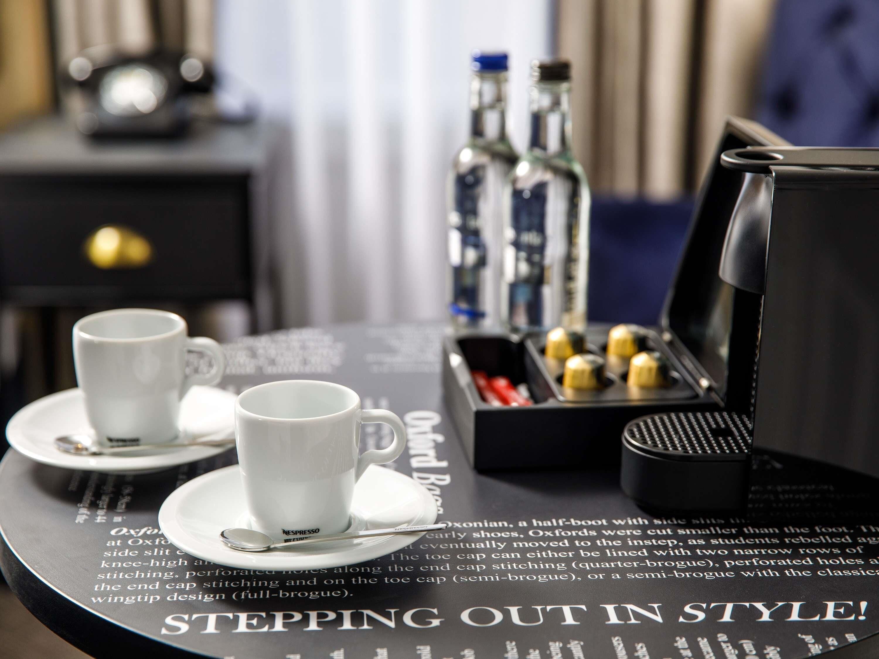 A hotel room with a coffee table featuring two white cups, a tray with condiments, and a coffee machine.