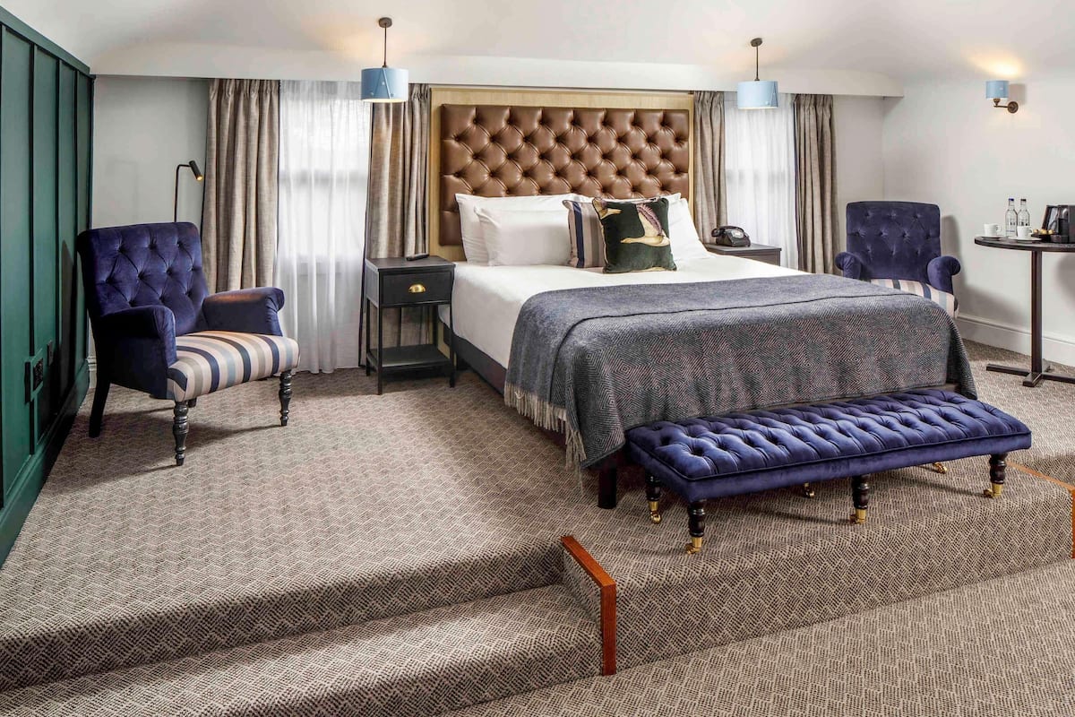 A hotel room with a tufted headboard, a large bed, a bench, a chair, a side table, and a lamp.