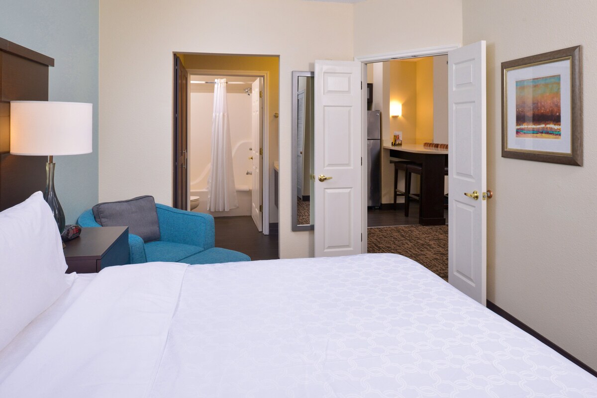 A hotel room with a bed, a blue armchair, a nightstand, a lamp, and a door leading to a bathroom and a dining area.