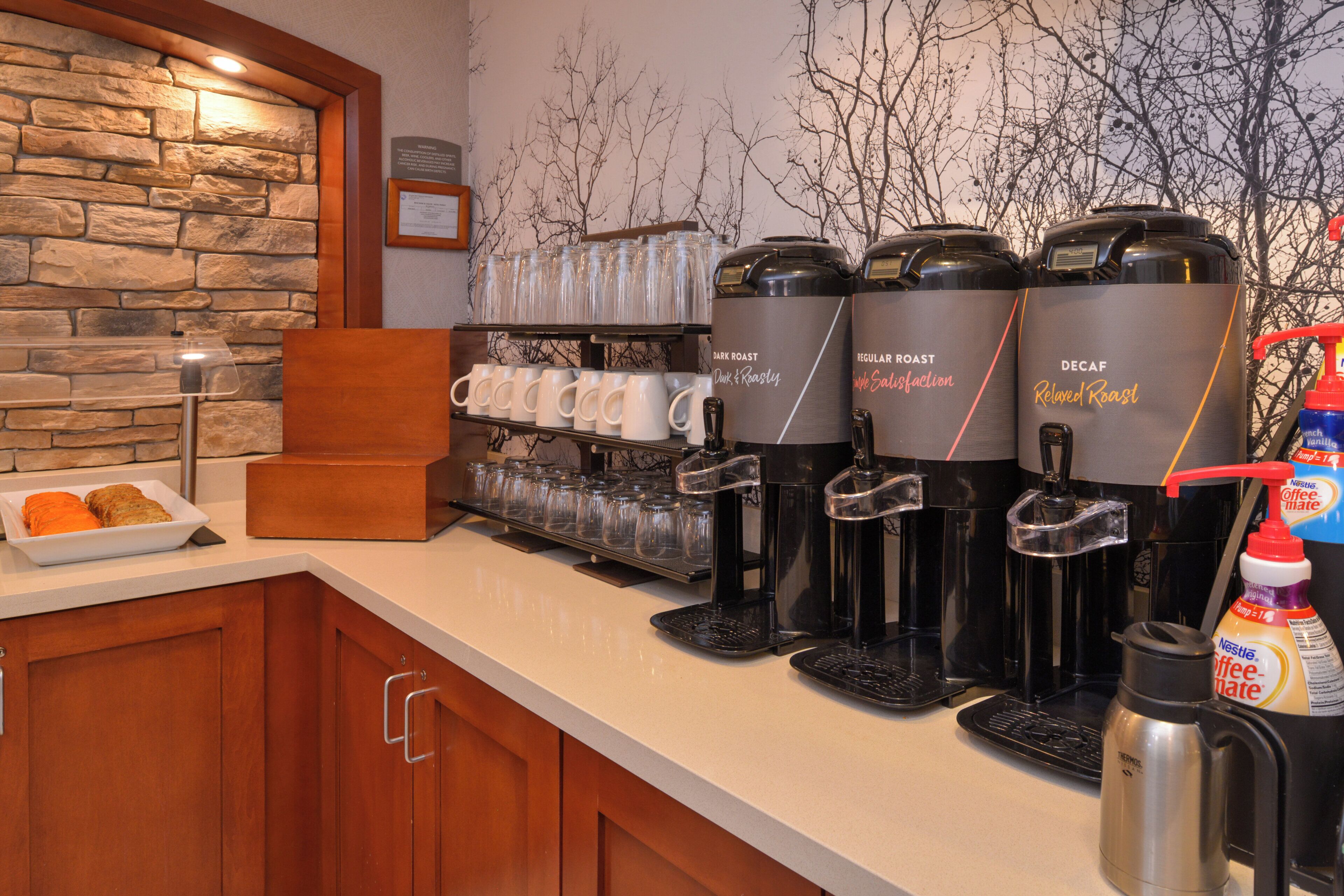 A coffee station with multiple coffee dispensers, a display of mugs, and a stone wall backdrop.