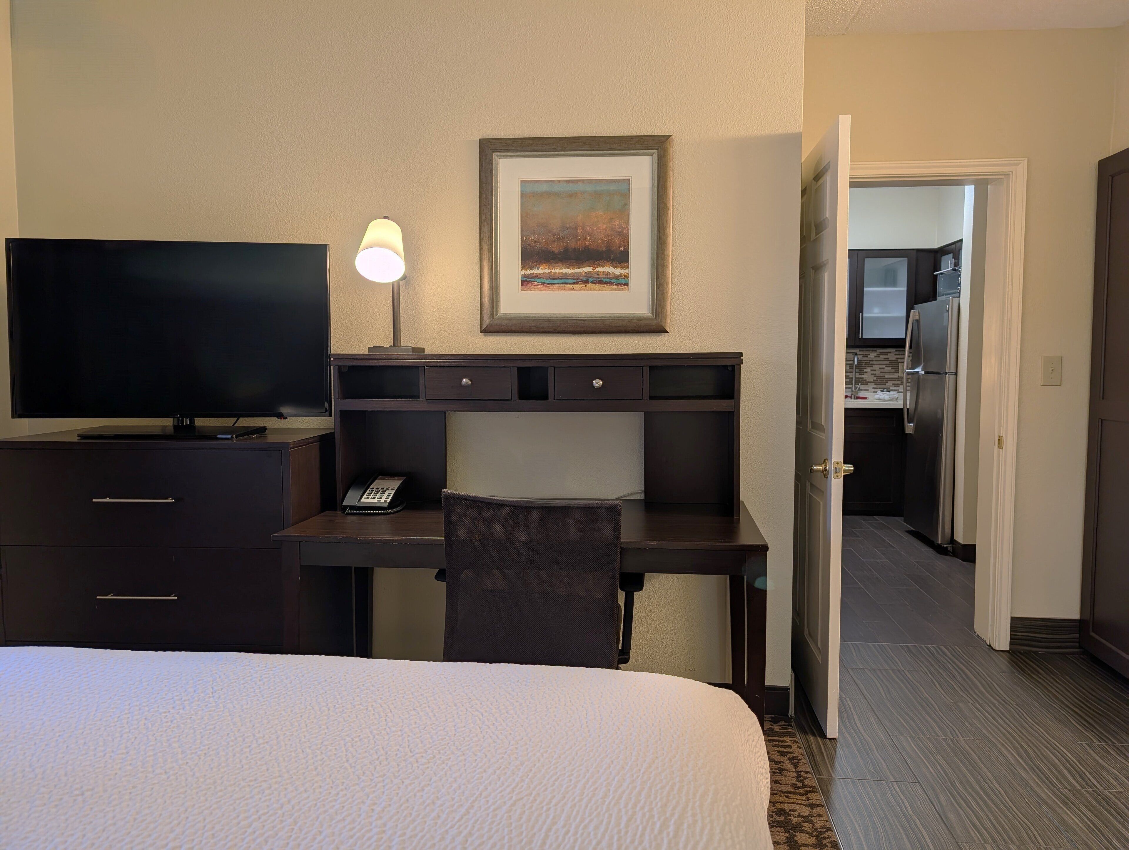 Ett hotellrum med en säng, ett skrivbord med en lampa, en tv och en tavla på väggen.
