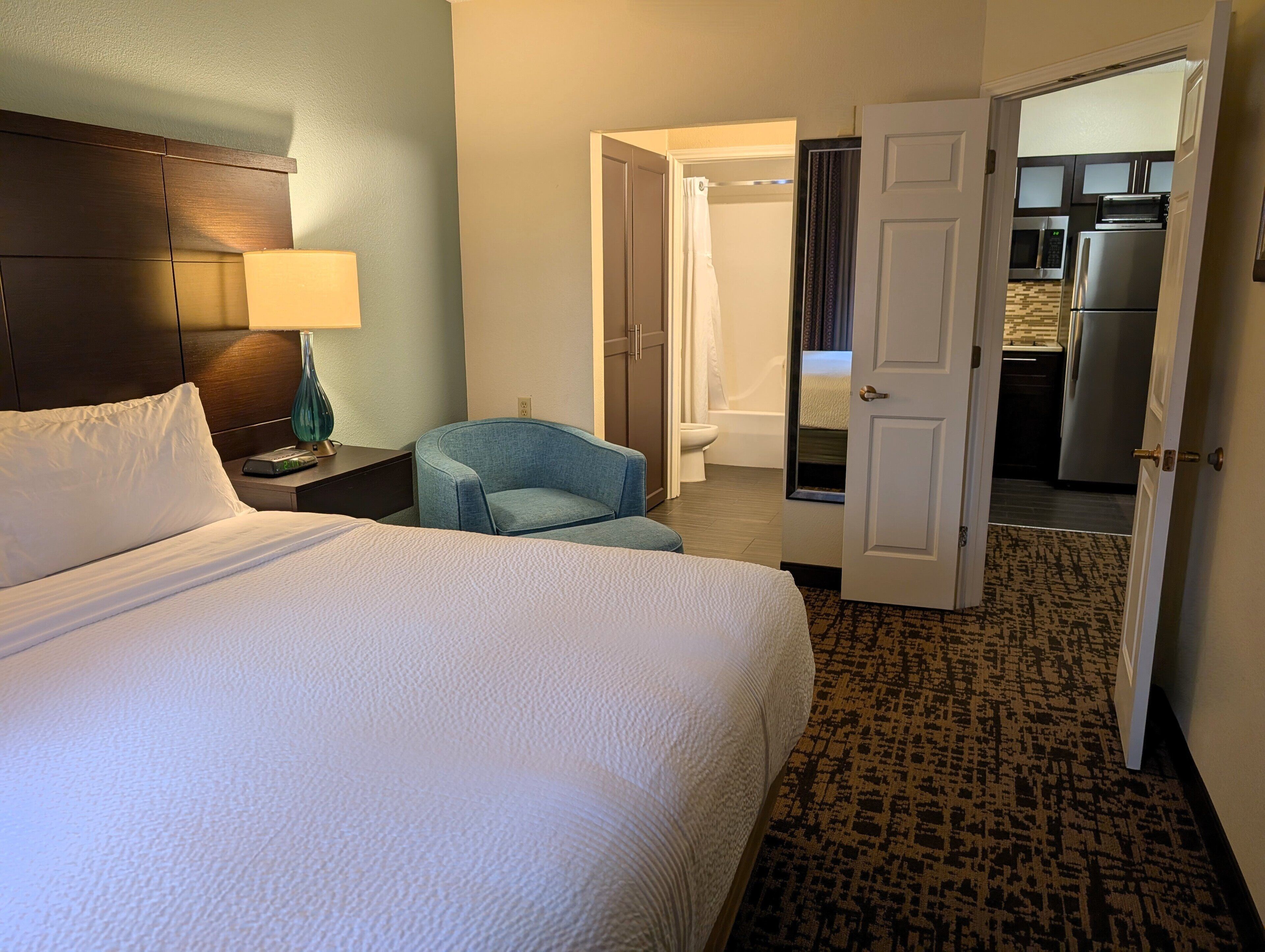 A hotel room with a bed, a chair, a nightstand with a lamp, and an open door leading to a bathroom and kitchen.