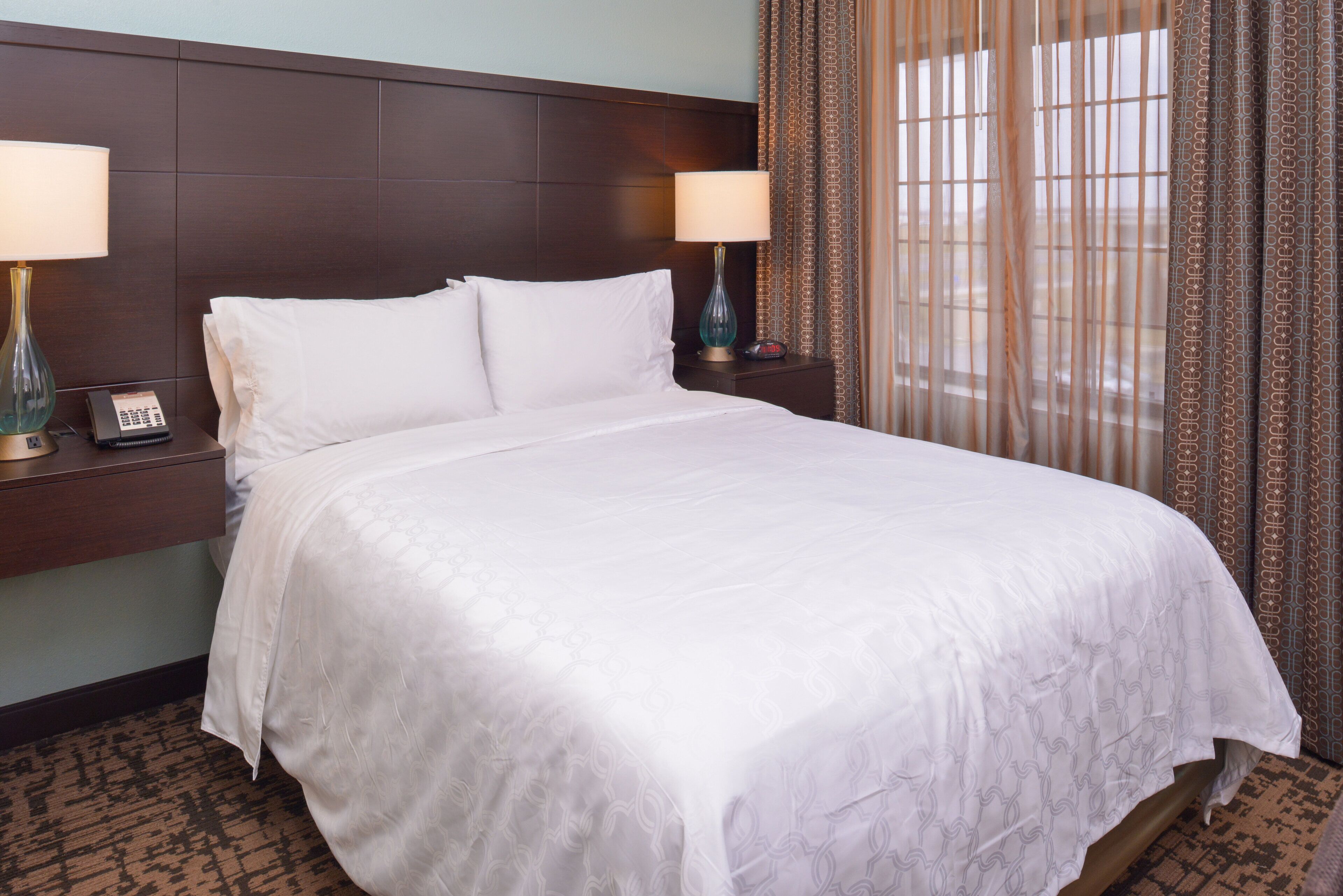 A neatly made bed with white linens, two bedside tables with lamps, and patterned curtains.