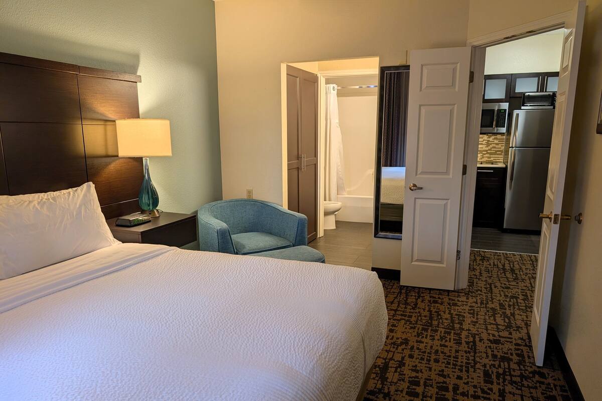 Une chambre d’hôtel avec un lit, une chaise, une table de chevet avec une lampe, et une porte ouverte donnant sur une salle de bain et une cuisine.