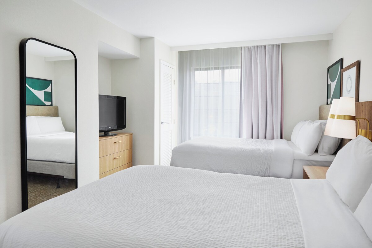 A hotel room with a large bed, a smaller bed, a TV on a wooden cabinet, and a window with curtains.