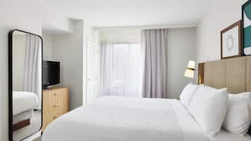 A hotel room with a large bed, a TV on a wooden dresser, a window with sheer curtains, and a framed picture on the wall.