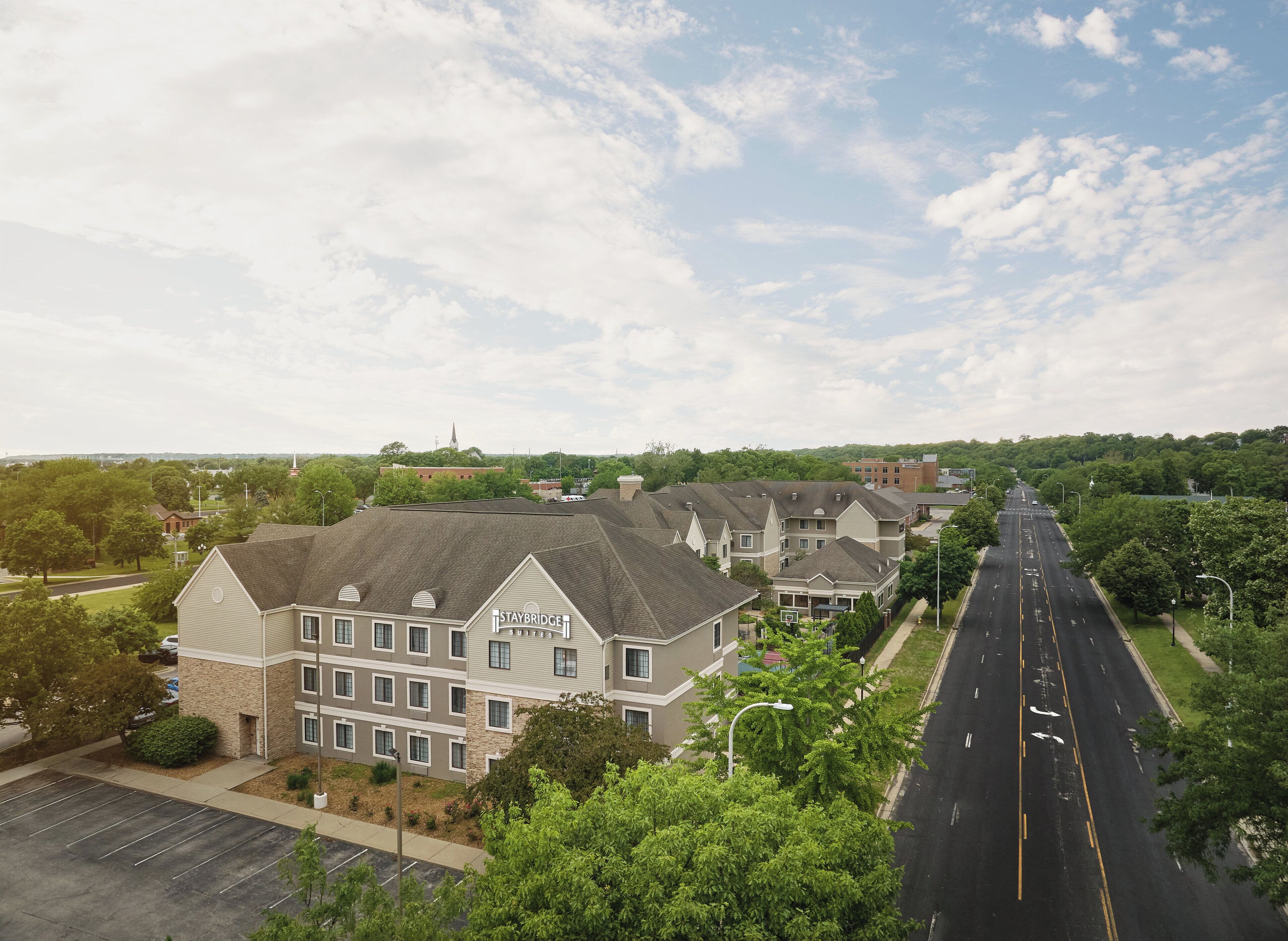 A Staybridge Suites hotel with a parking lot, a road with a median, and surrounding greenery.