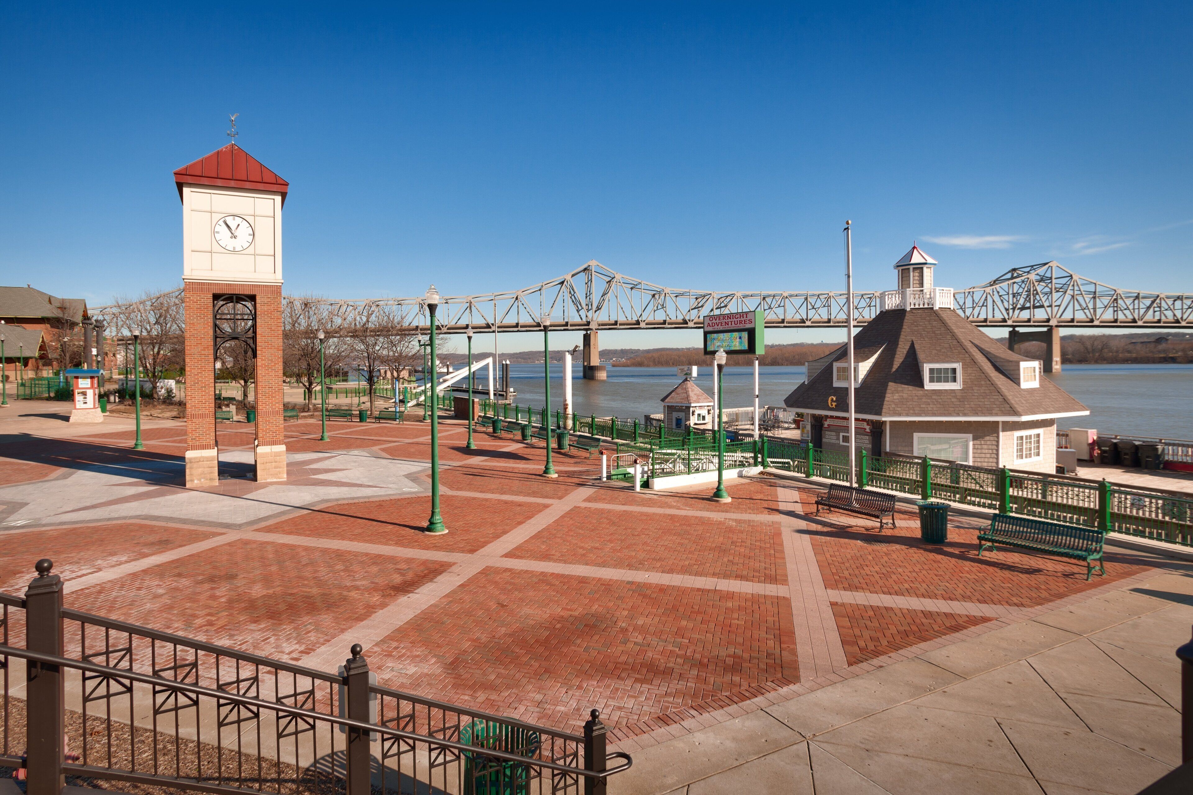 A riverside walkway with a clock tower, benches, and a bridge in the background.