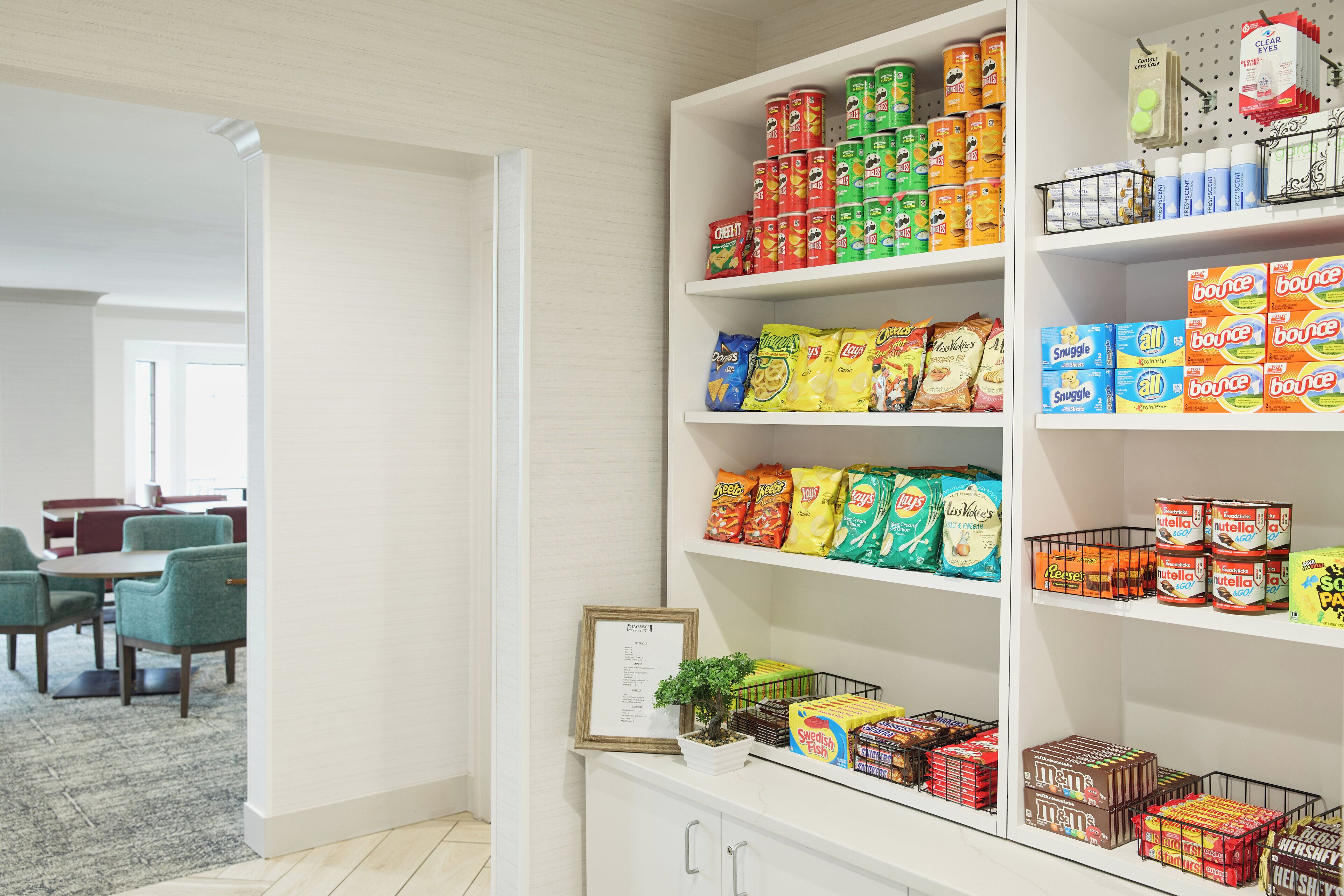 A pantry stocked with various snacks and beverages, a framed certificate on the counter, and a small potted plant.