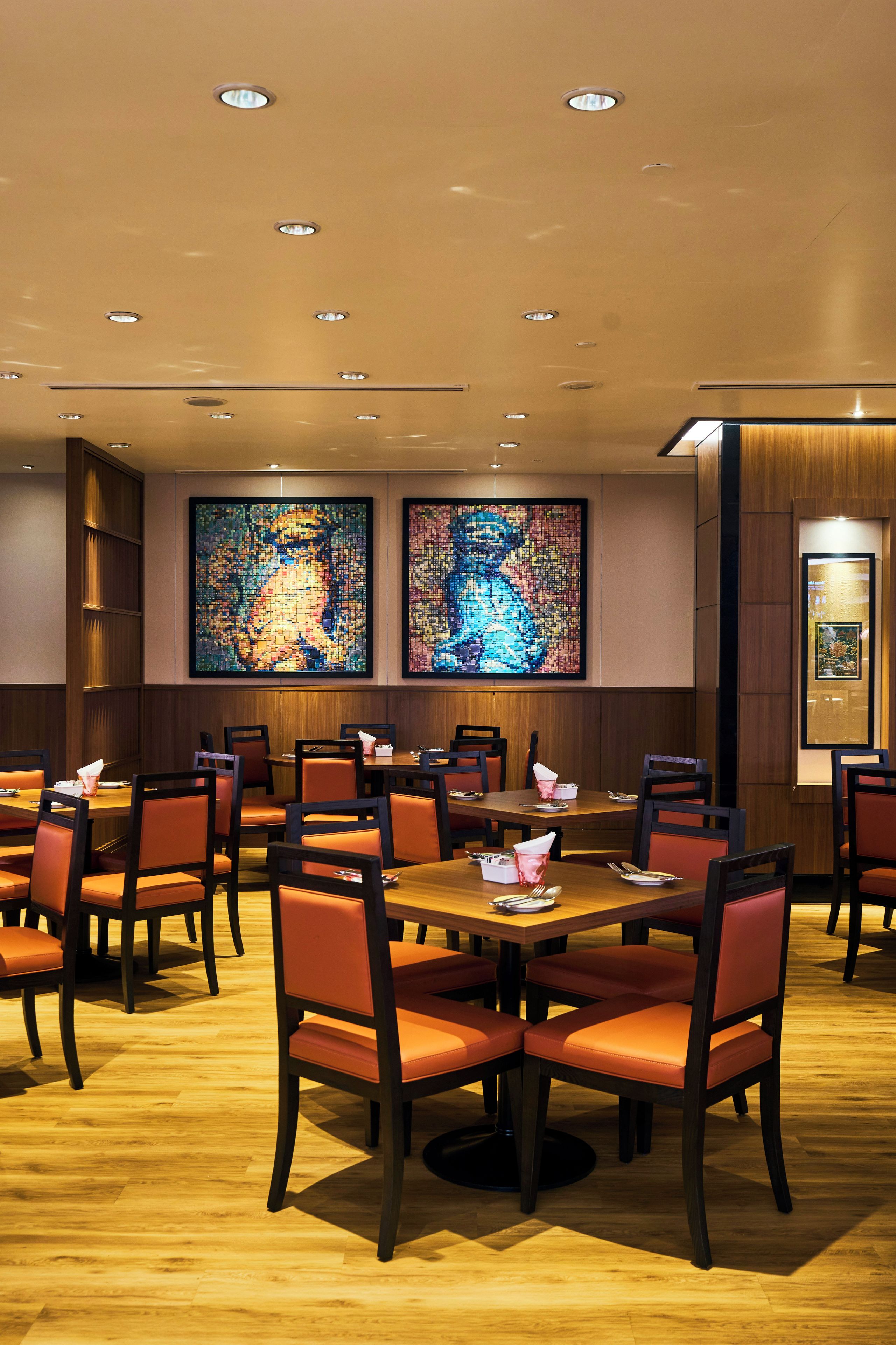 A restaurant with wooden tables and chairs, recessed ceiling lights, and two large mosaic artworks on the walls.