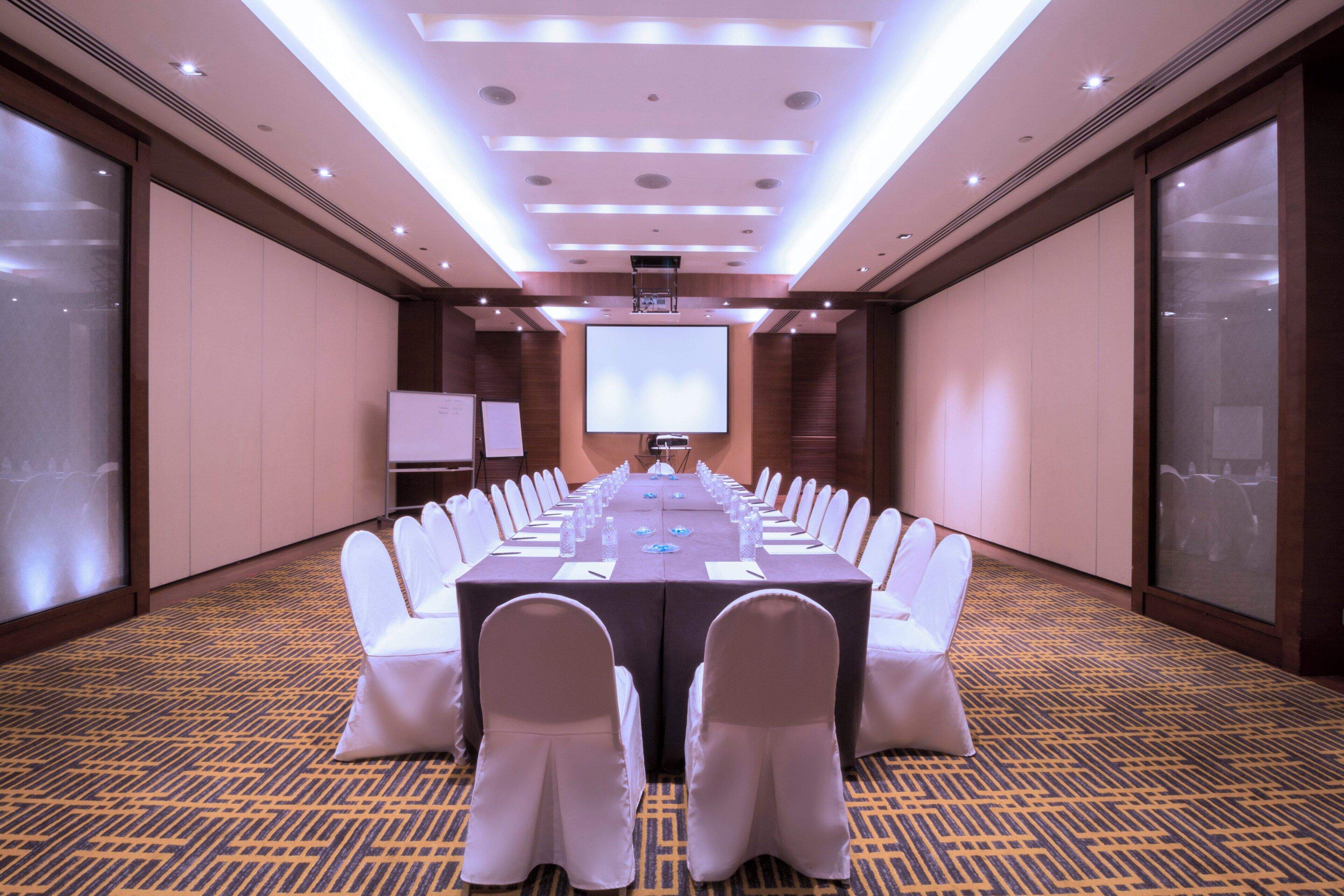 Un salón de conferencias con una larga mesa, sillas blancas y una pantalla grande al frente.