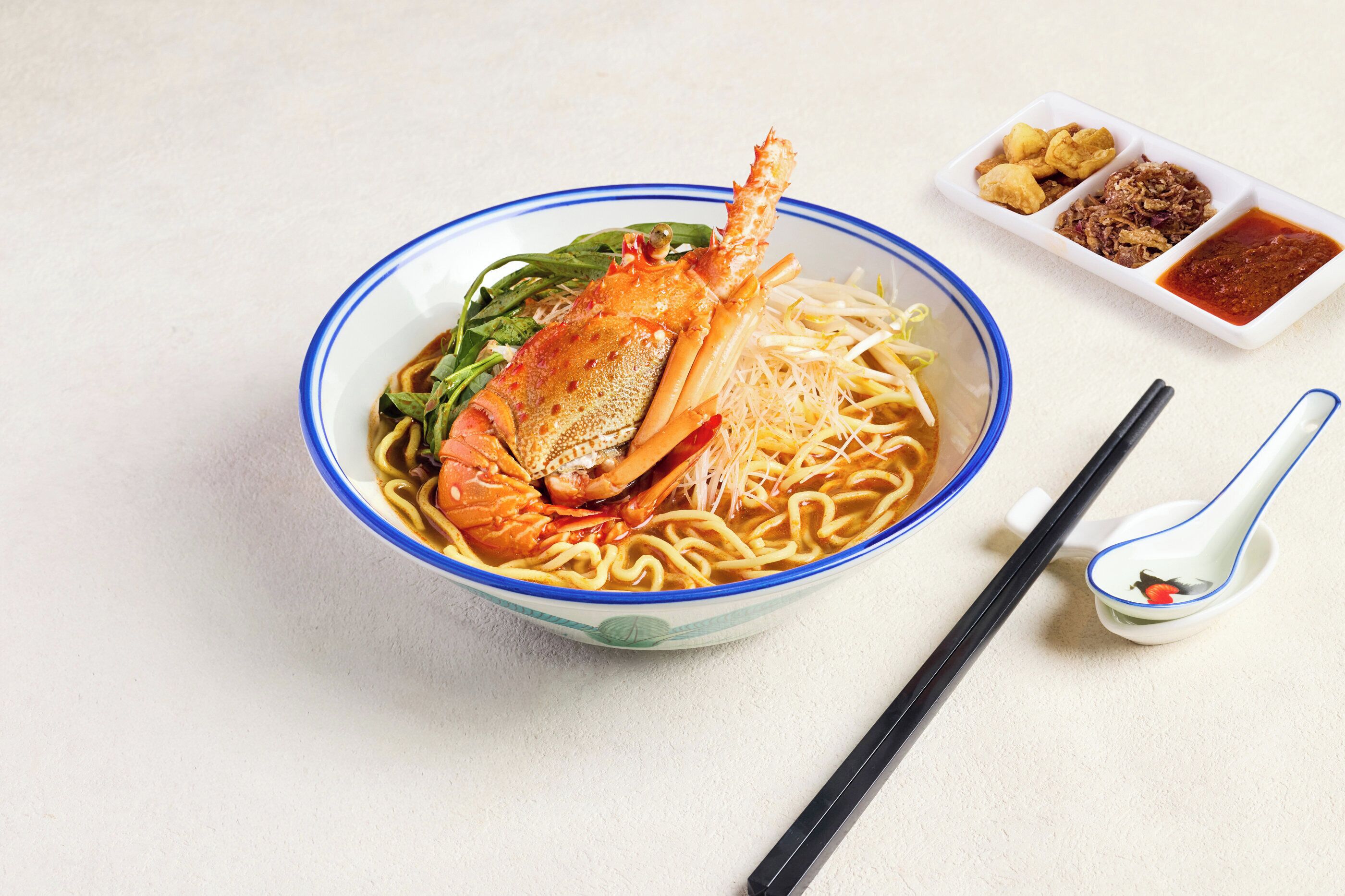 Un tazón de fideos con cangrejo, verduras verdes y dos pequeños recipientes de condimentos.