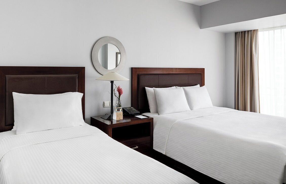 Een hotelkamer met twee bedden, een nachtkastje met een lamp, een spiegel en een raam met gordijnen.