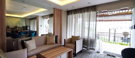 A modern hotel room with a sofa, armchairs, a wooden coffee table, and a balcony with a view.