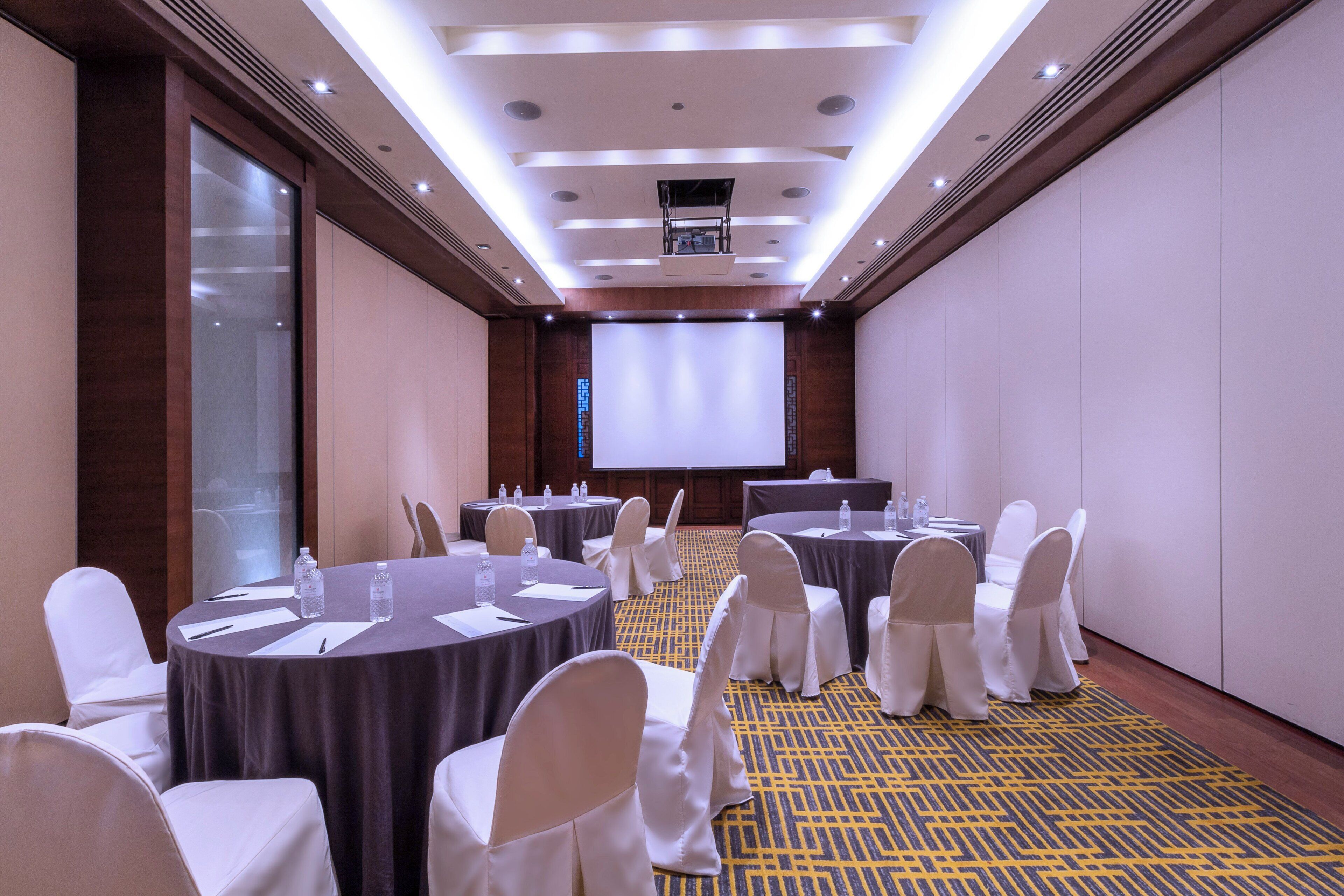 A conference room with round tables, white chairs, and a large screen at the front.