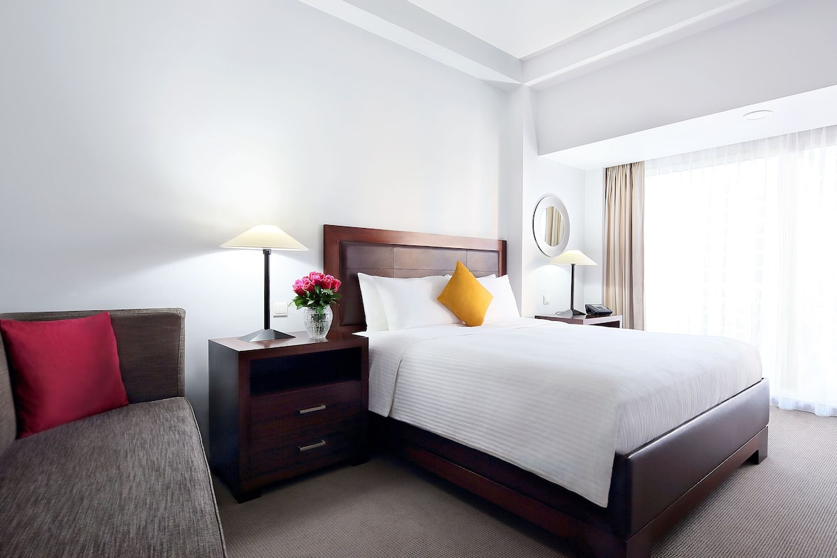 Une chambre d’hôtel avec un grand lit, une table de chevet avec une lampe, une commode, une chaise et une fenêtre avec des rideaux.