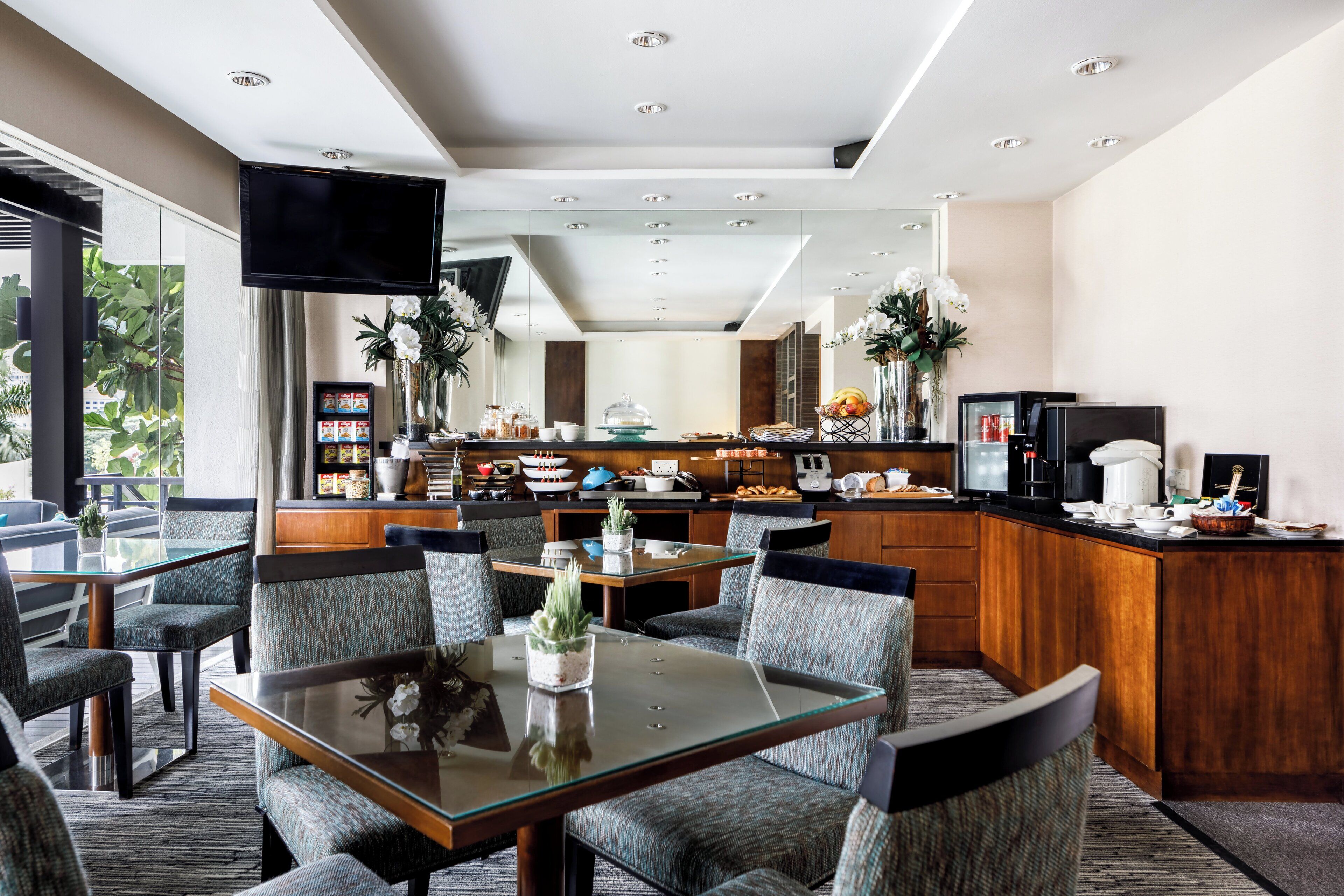 A modern hotel dining area with a glass table, cushioned chairs, and a kitchen in the background.