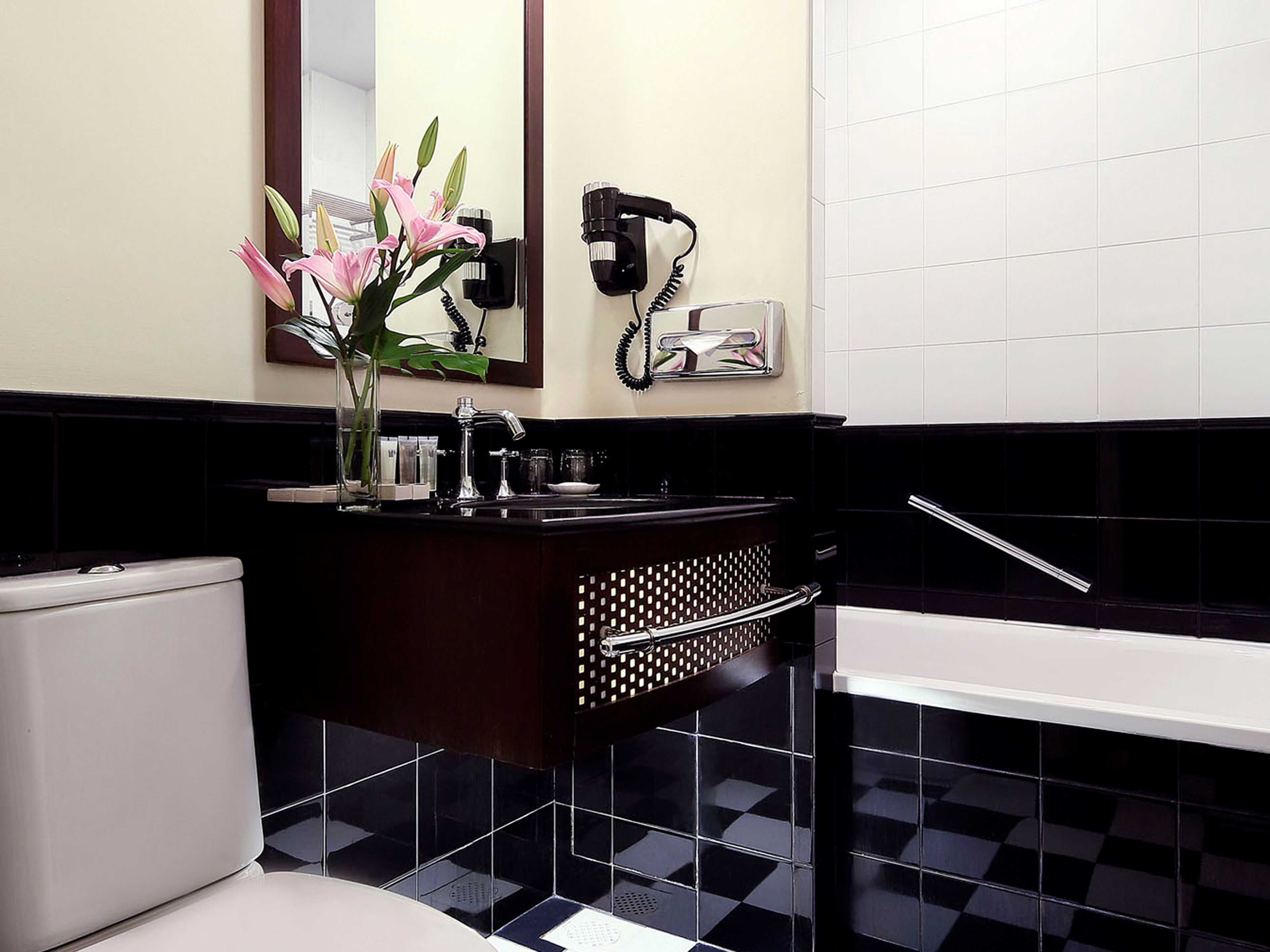 Un baño con piso blanco y negro a cuadros, un inodoro blanco, un lavamanos con espejo y un florero con flores.