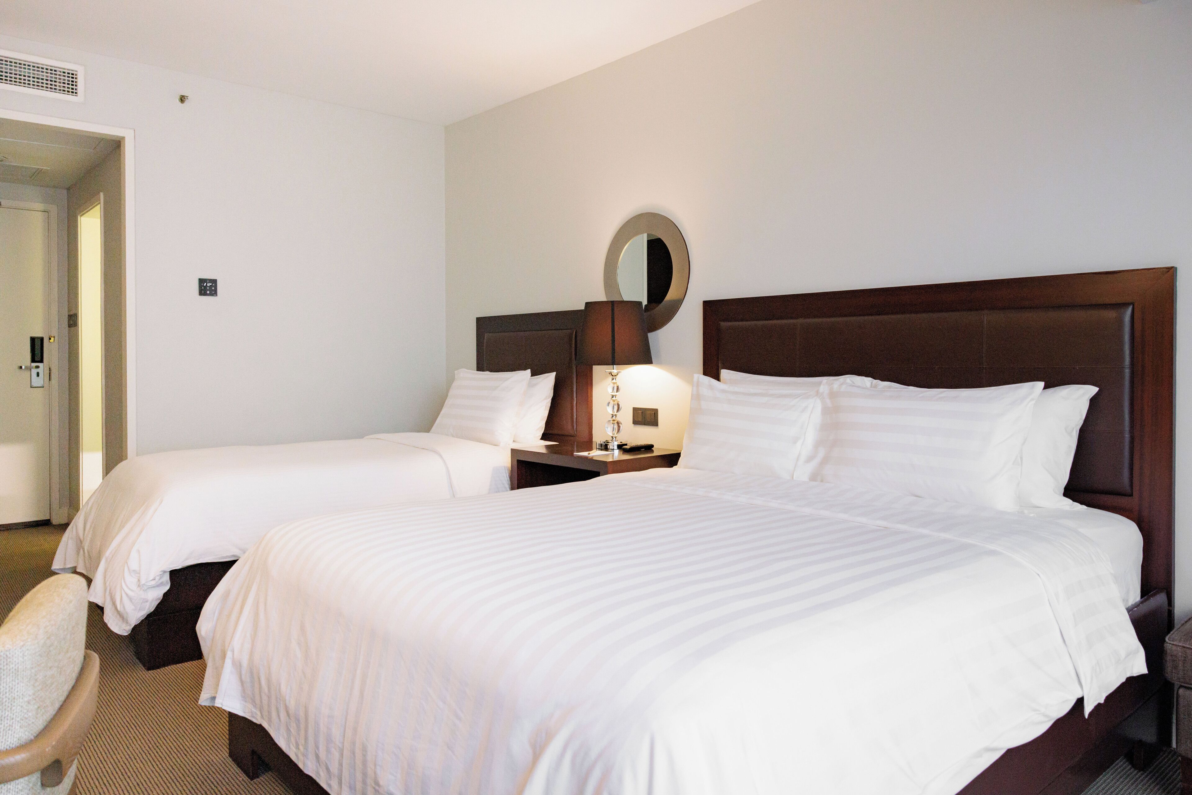 A hotel room with two beds, a bedside table with a lamp, a mirror, and a chair.