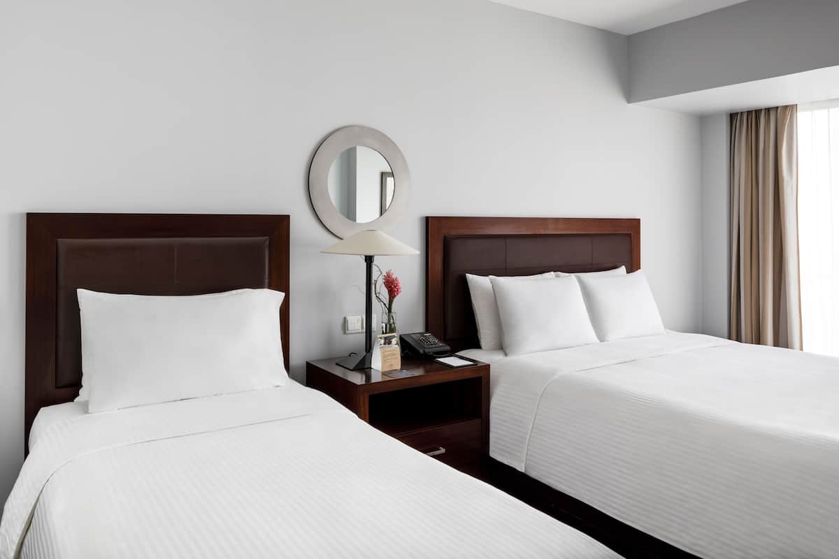 Une chambre d’hôtel avec deux lits, une table de chevet avec une lampe, un miroir et une fenêtre avec des rideaux.