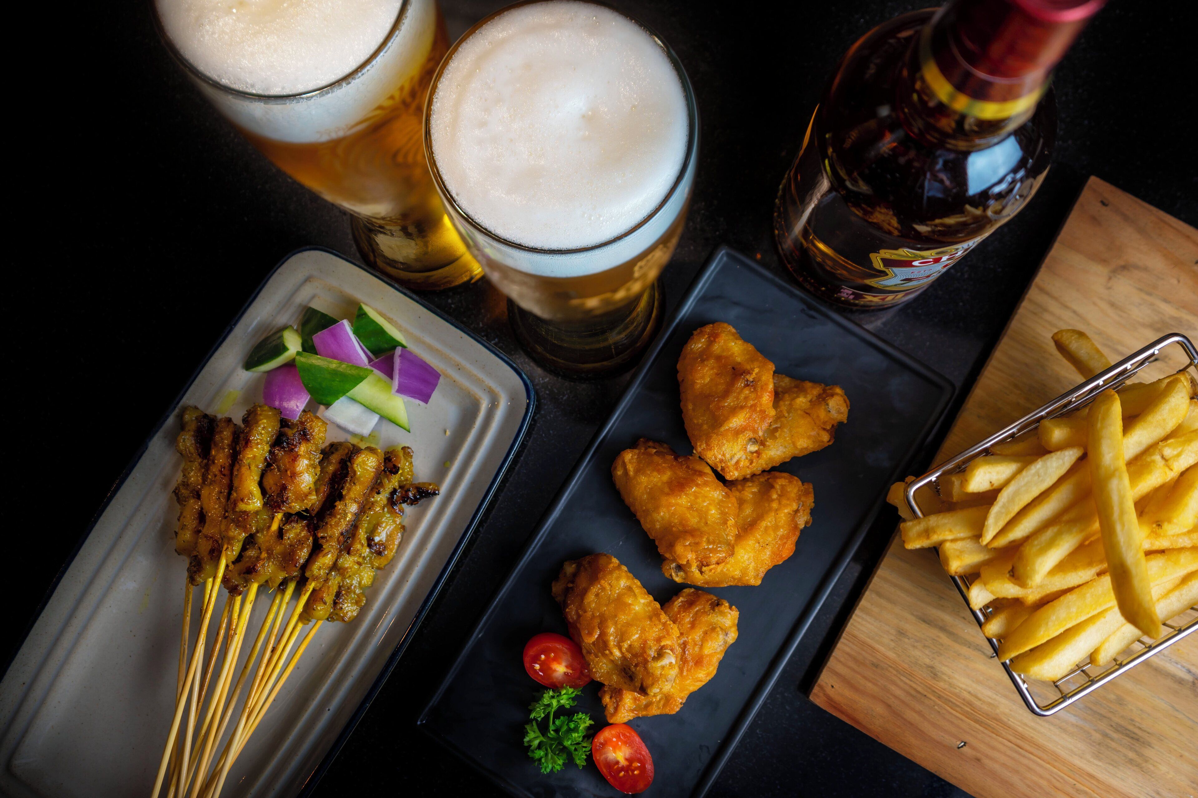 A meal with satay, fried chicken, and fries, accompanied by two glasses of beer and a bottle of sauce.