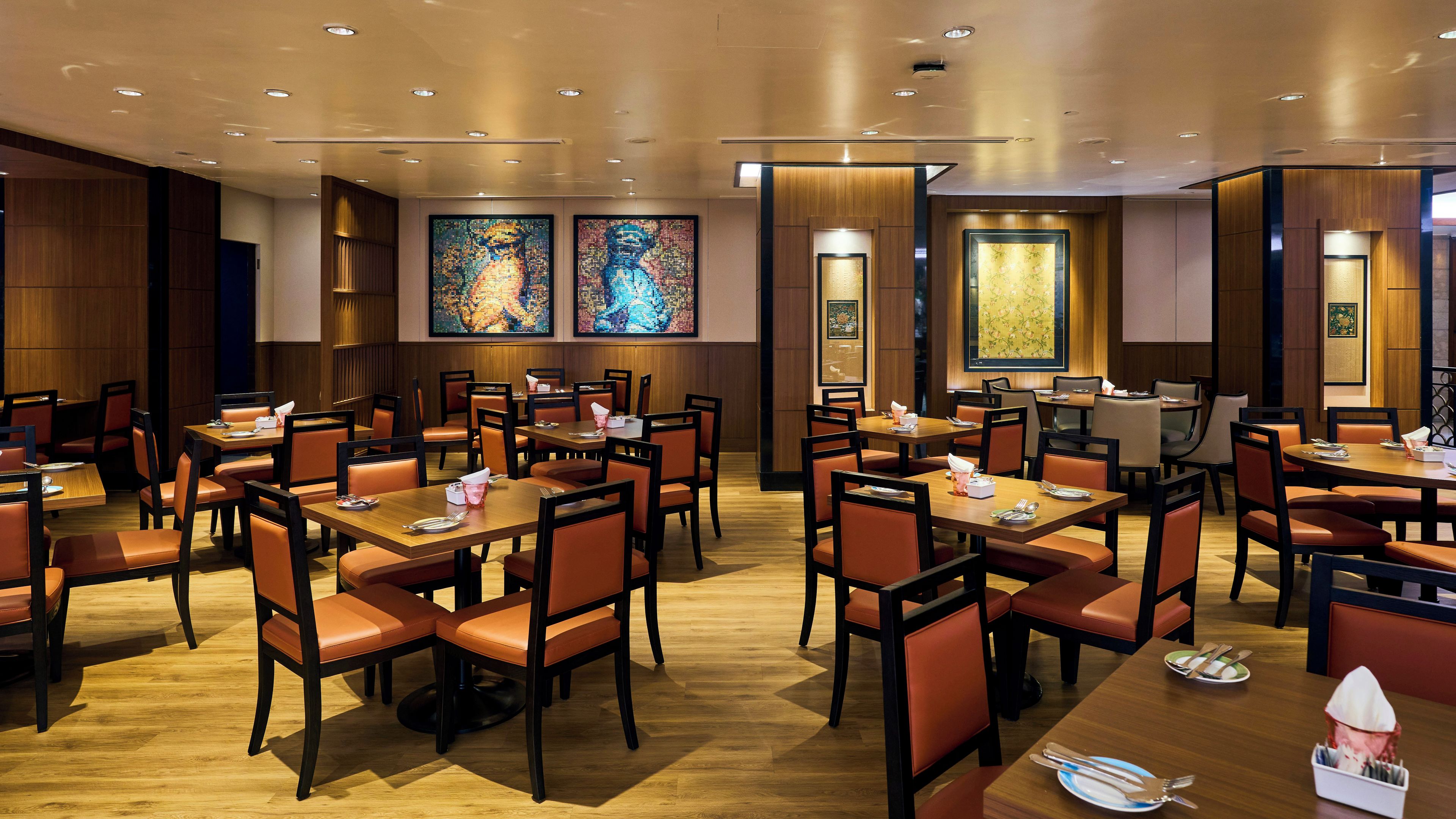 A hotel dining area with multiple tables and chairs, wooden flooring, and artwork on the walls.