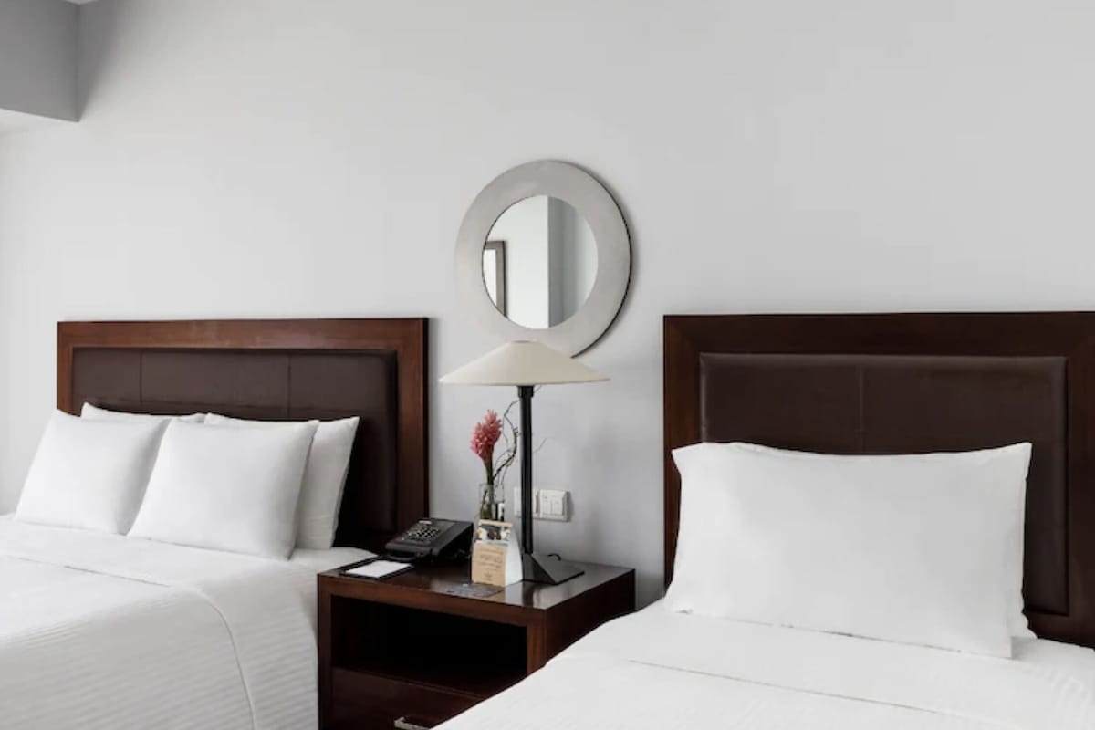 Una habitación de hotel con dos camas, una mesita de noche con lámpara, un espejo y una ventana con cortinas.