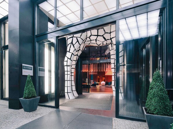 Een moderne hotelentree met een unieke geometrische boog, een receptie met rood verlicht interieur en twee potplanten op de vloer.