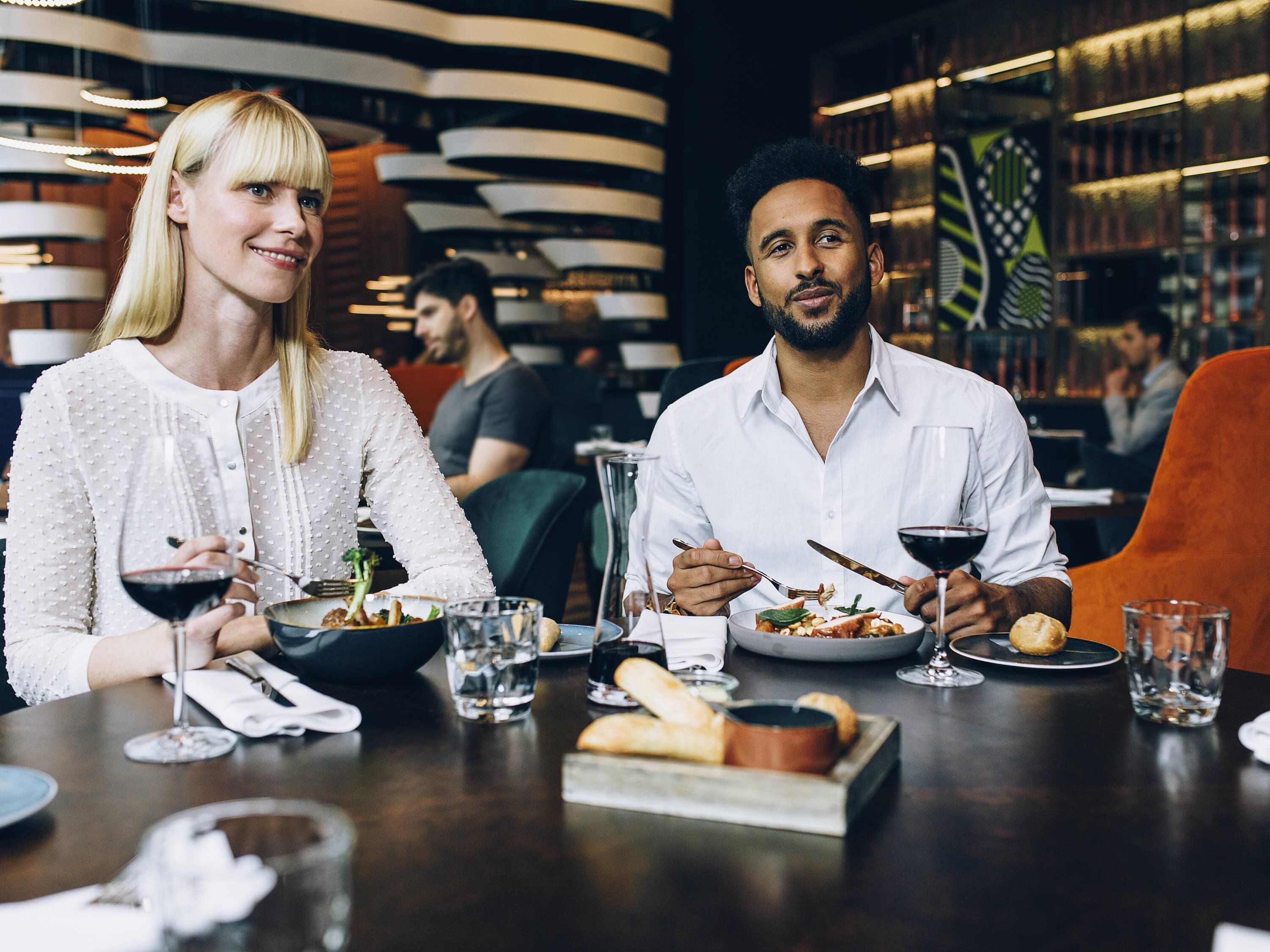 A couple dining in a modern restaurant with a contemporary interior, featuring a dark wooden table and orange chairs.