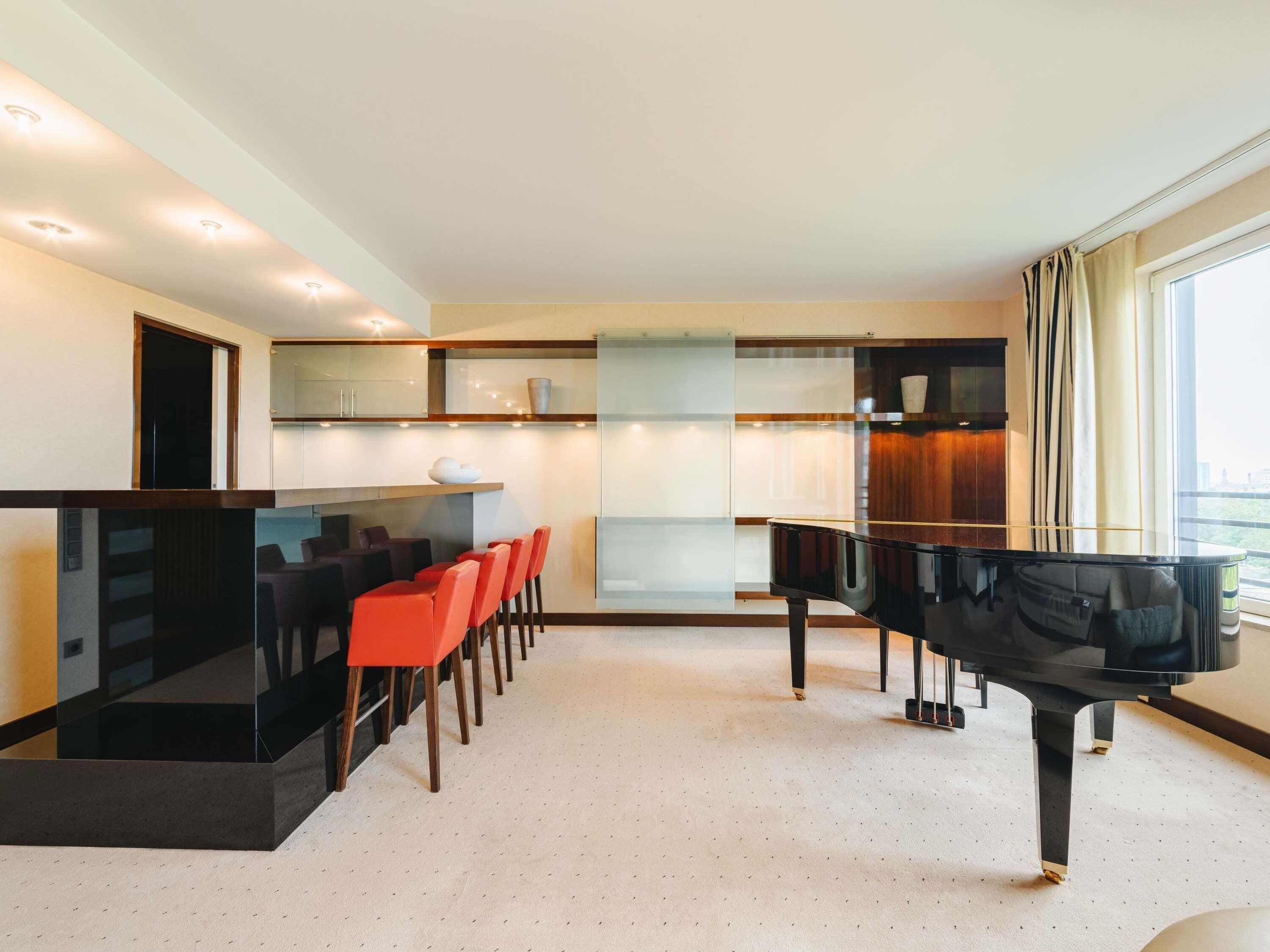 Ein modernes Wohnzimmer mit einem Klavier, einem Essbereich mit roten Stühlen, einer Küche mit Glastüren und einem großen Fenster mit Vorhängen.