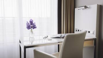 Une chambre d’hôtel moderne avec un bureau en verre, un ordinateur portable, un vase rempli de fleurs violettes et un grand miroir au mur.